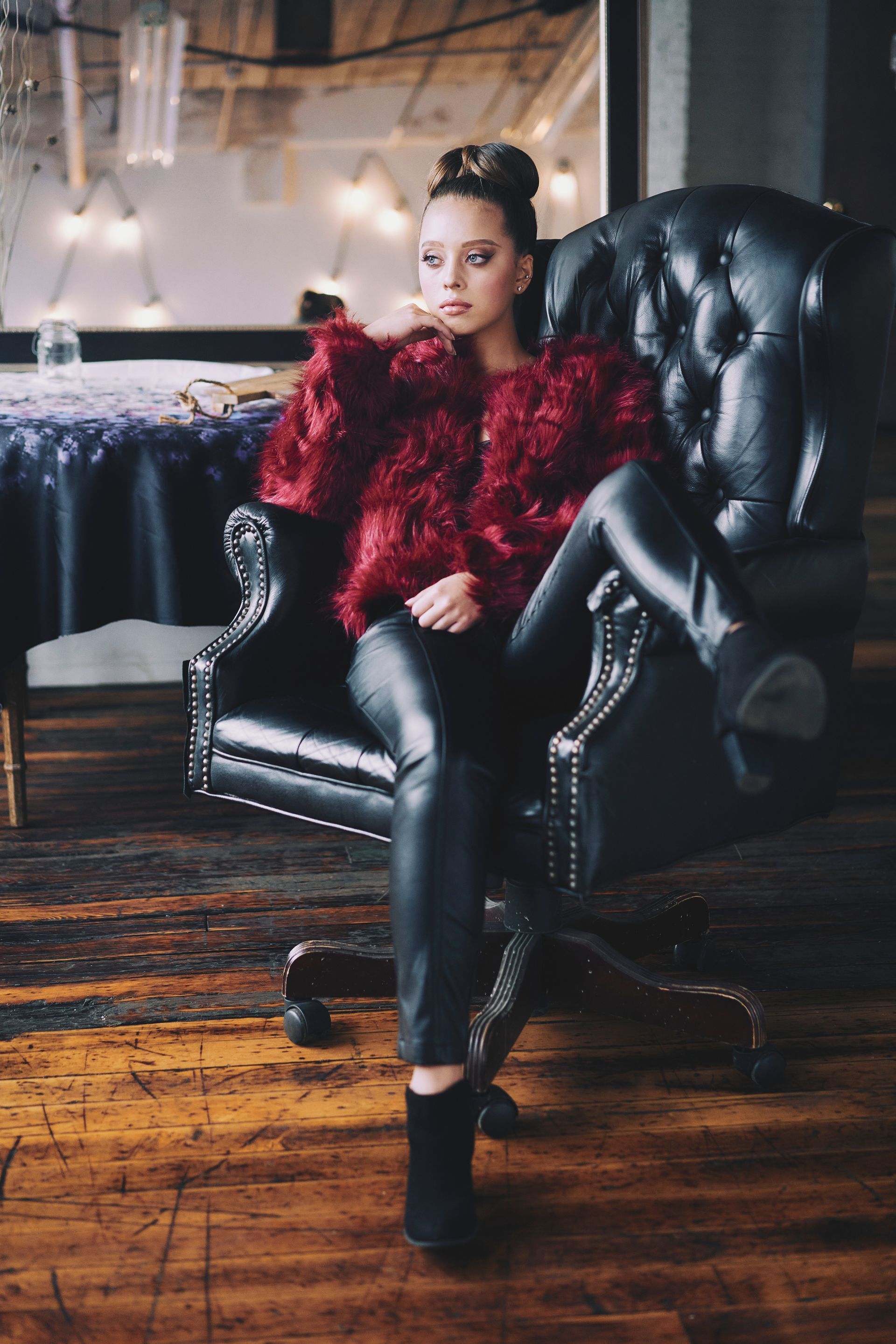 Woman in red fuzzy jacket and black leather pants sits in a black tufted armchair.