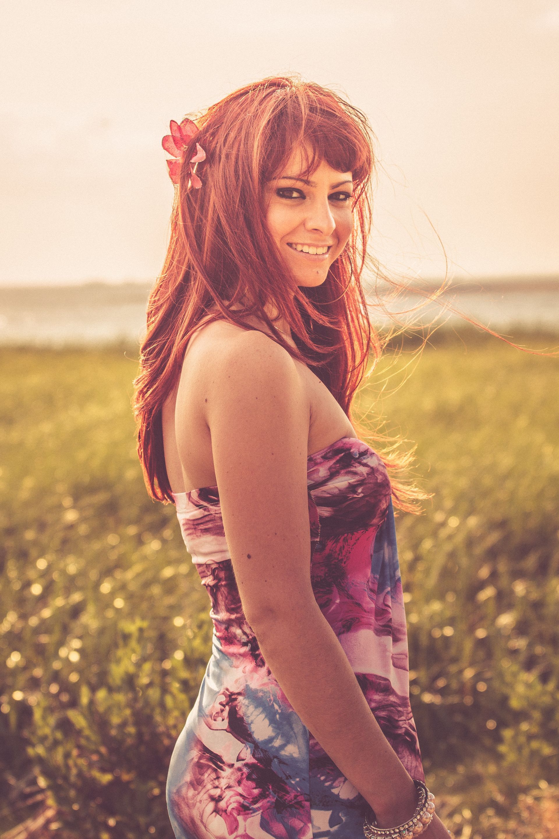 Woman with red hair and flower smiling in field, wearing floral dress.