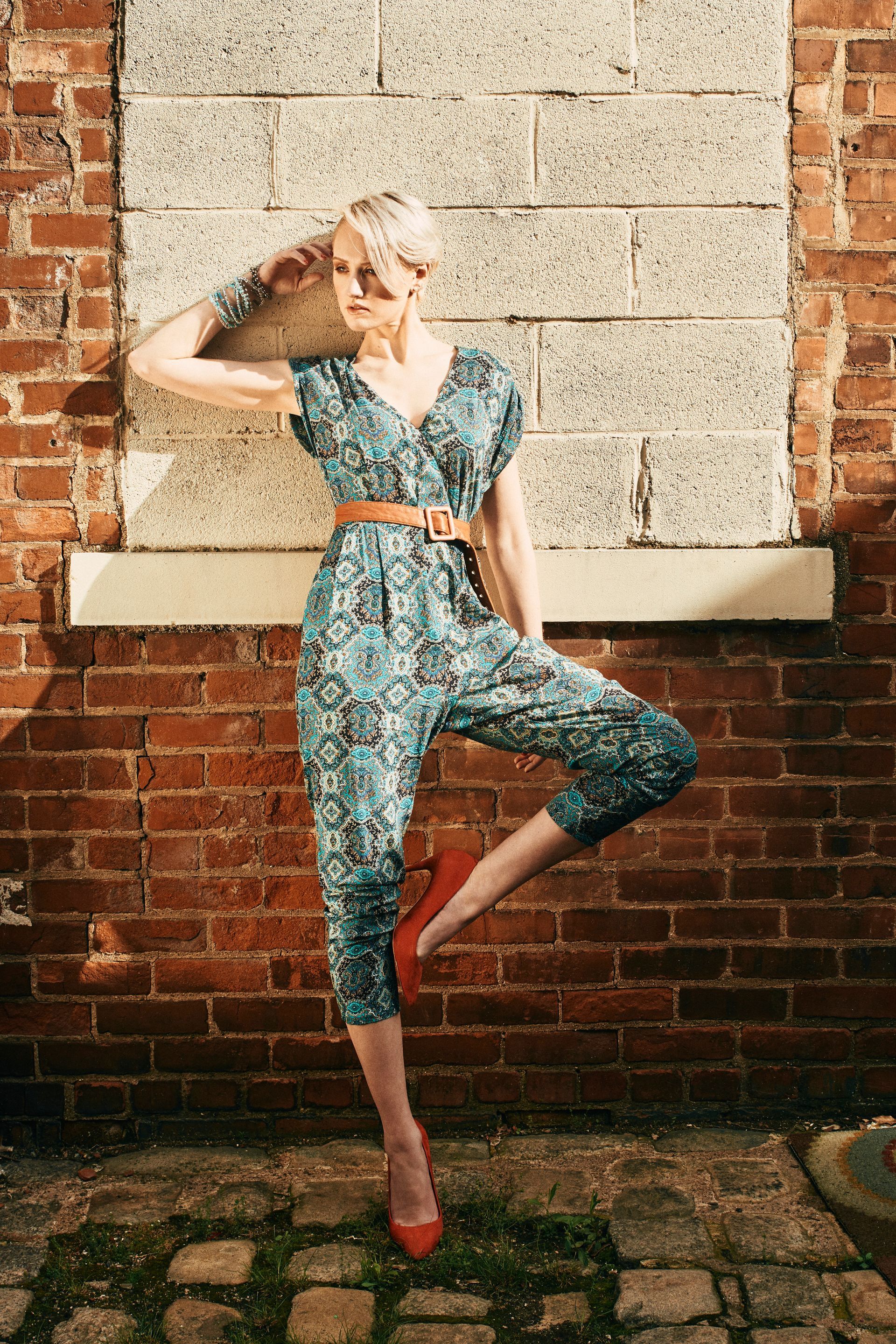 Woman in blue jumpsuit and red heels poses by brick wall.