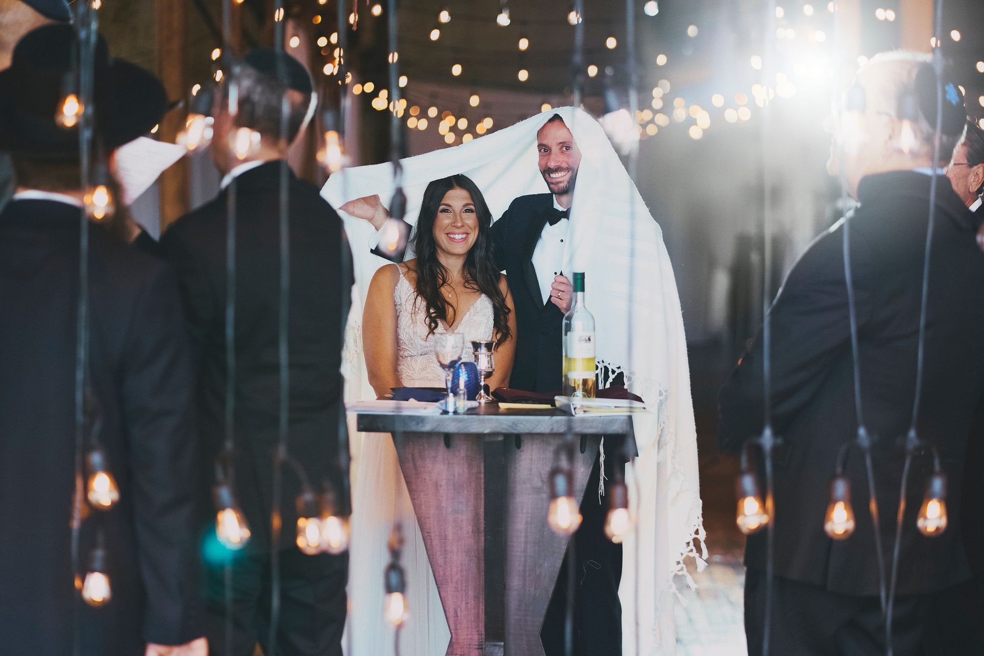 Bride and groom under a cloth, flanked by men in suits, celebrating with drinks, lights.