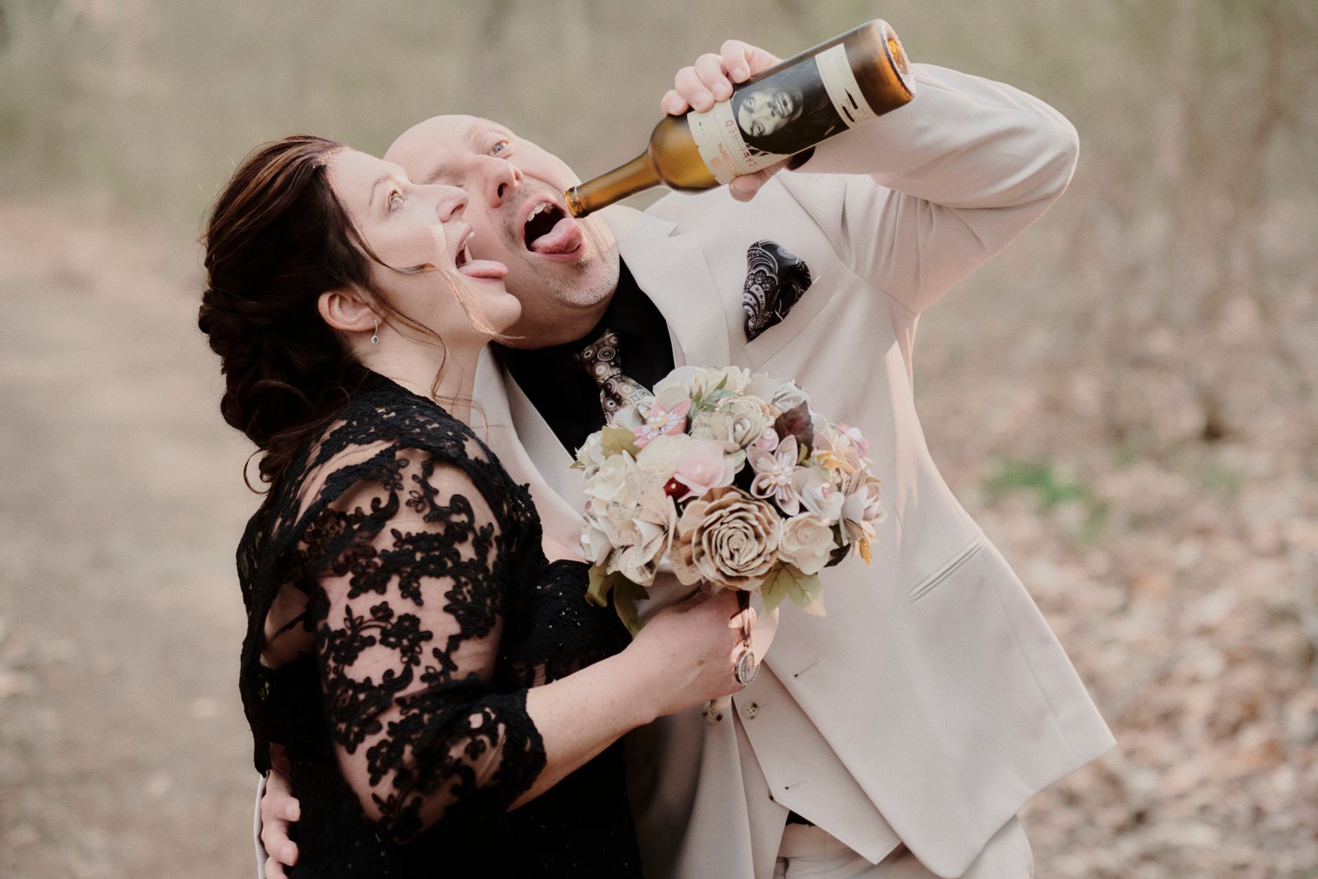 Newlyweds drinking wine from the bottle, mouths open, outdoors. Woman in black lace, man in beige suit, bouquet.