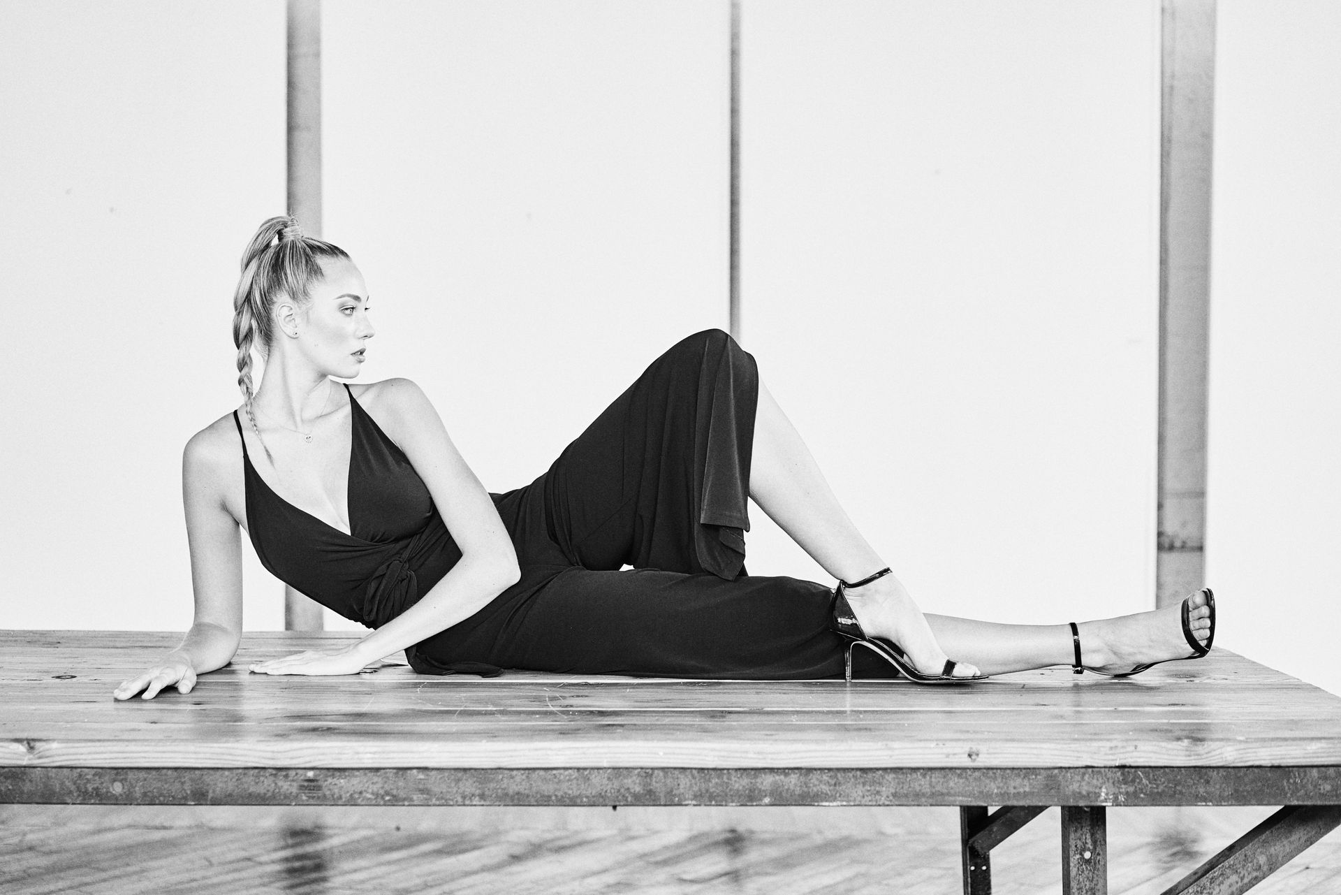 Woman in black jumpsuit reclines on a table, looking away. She has blonde hair and is wearing heels.