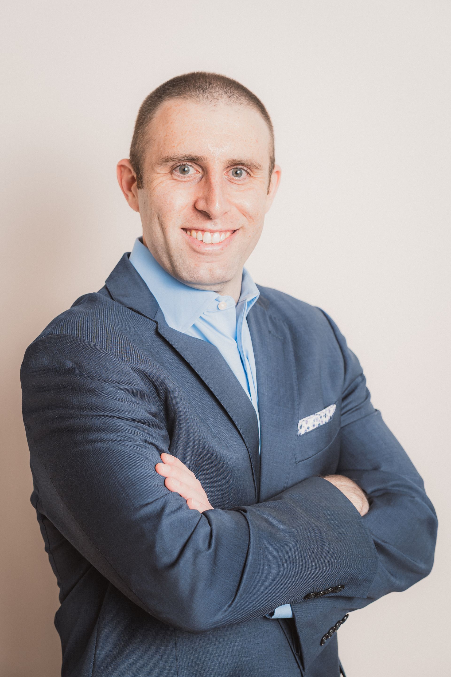 Man with arms crossed smiling, wearing blue blazer and shirt, against a light background.