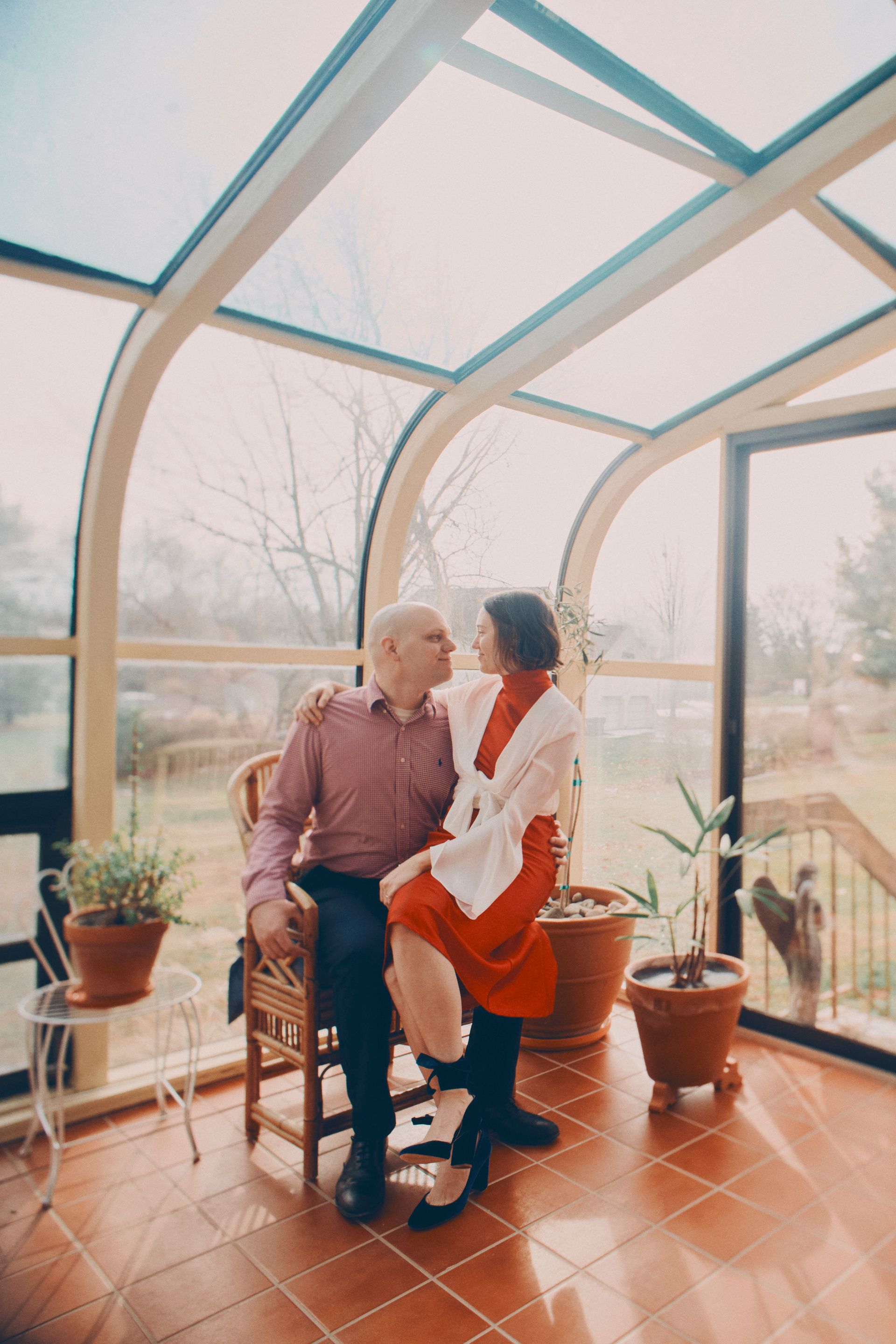 Couple in sunroom, man seated, woman perched on his lap, looking at each other. Red and white clothing, sunny interior.