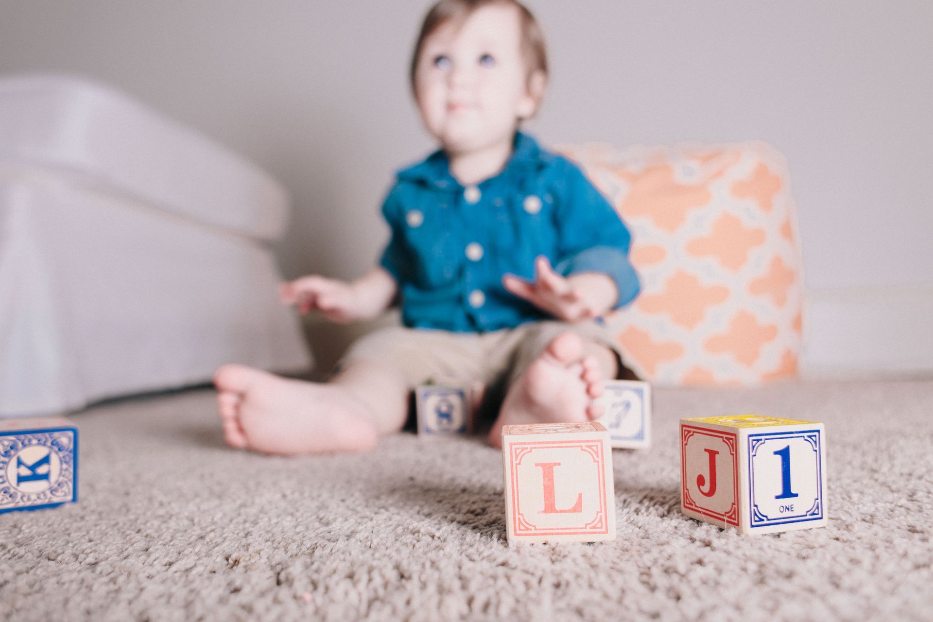 Baby playing with alphabet blocks on a carpeted floor, looking up with interest.