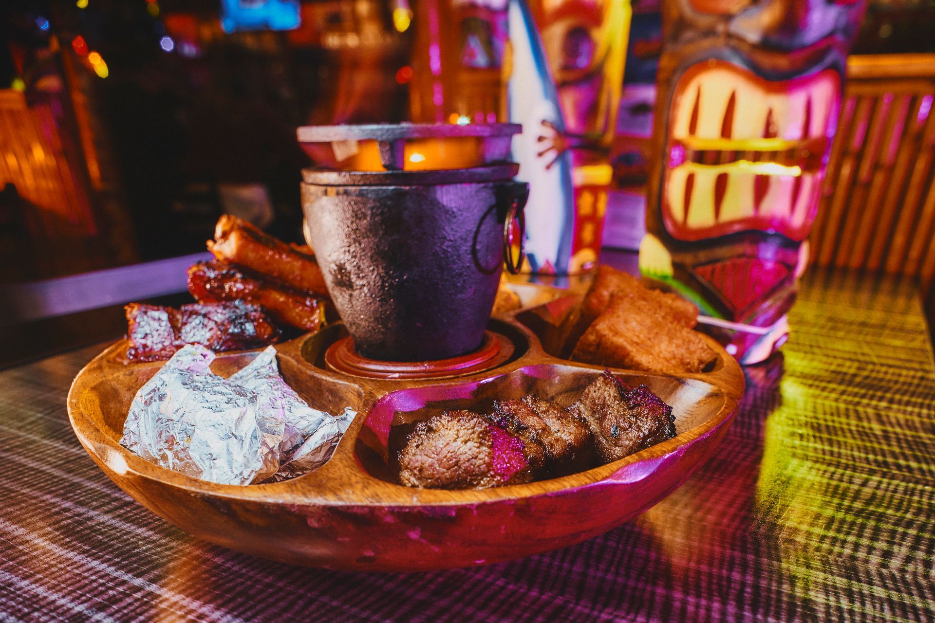 Wooden platter with various meats, bread, and a central pot of fire in a tiki bar.