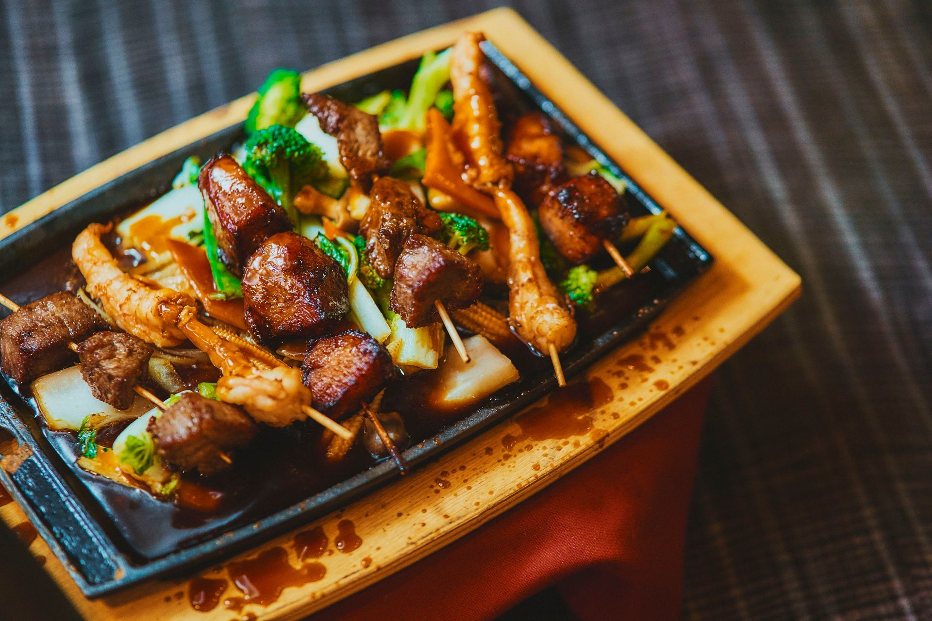 Skewered meat and vegetables on a sizzling black plate with sauce, served on a wooden tray.
