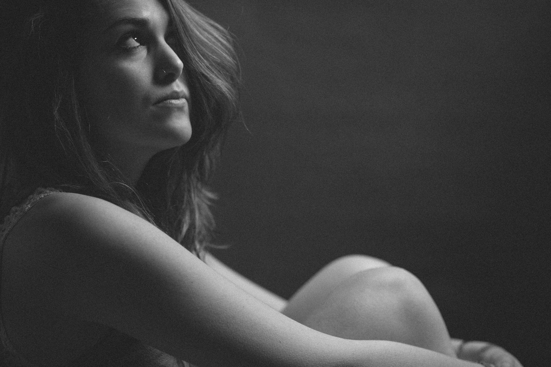 Woman in black and white, looking up pensively, arms around her legs, dark background.