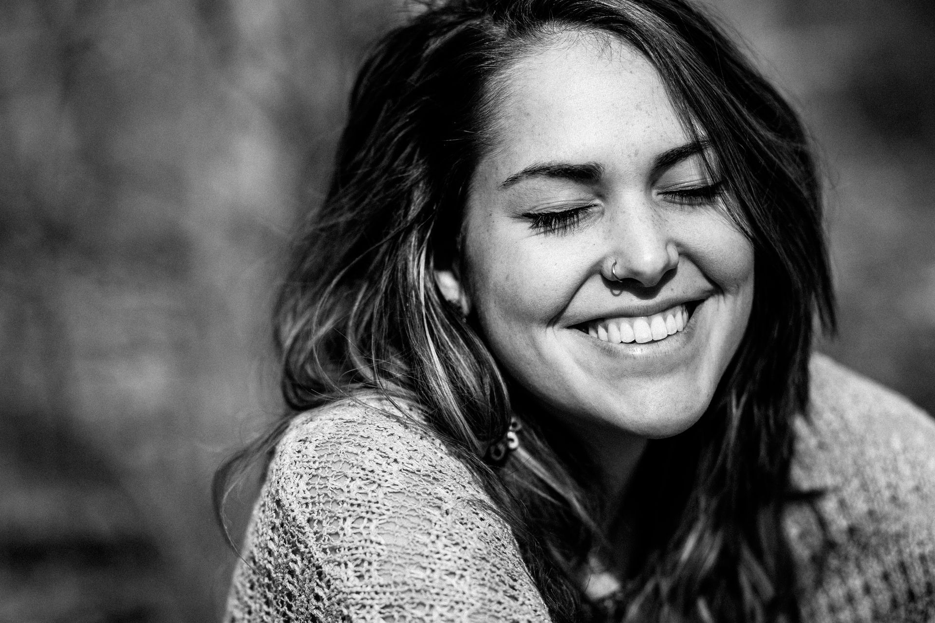 Woman with closed eyes and broad smile, nose ring, textured sweater, in black and white.