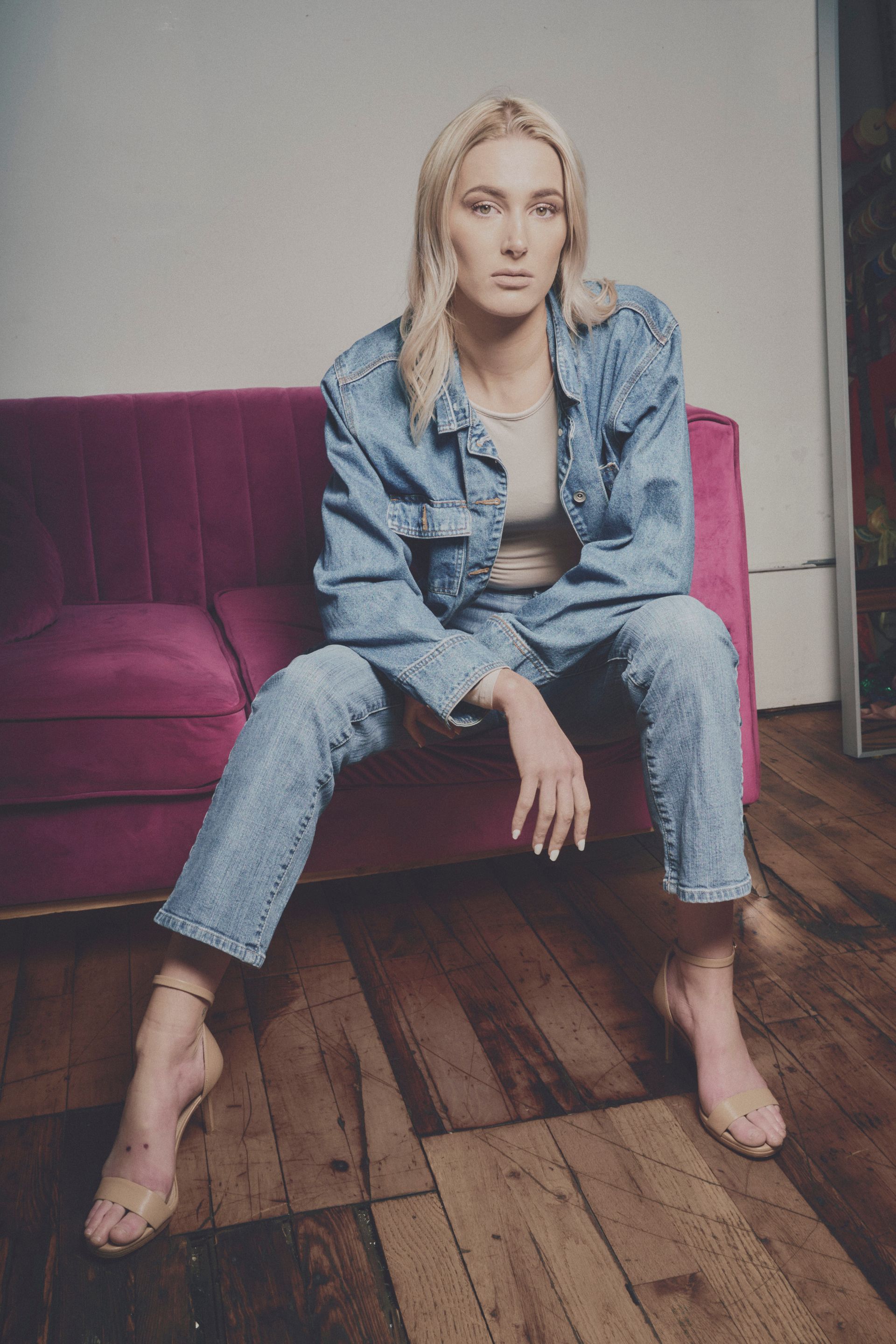 Blonde woman in denim on a magenta couch, wearing heels, looking at the camera.