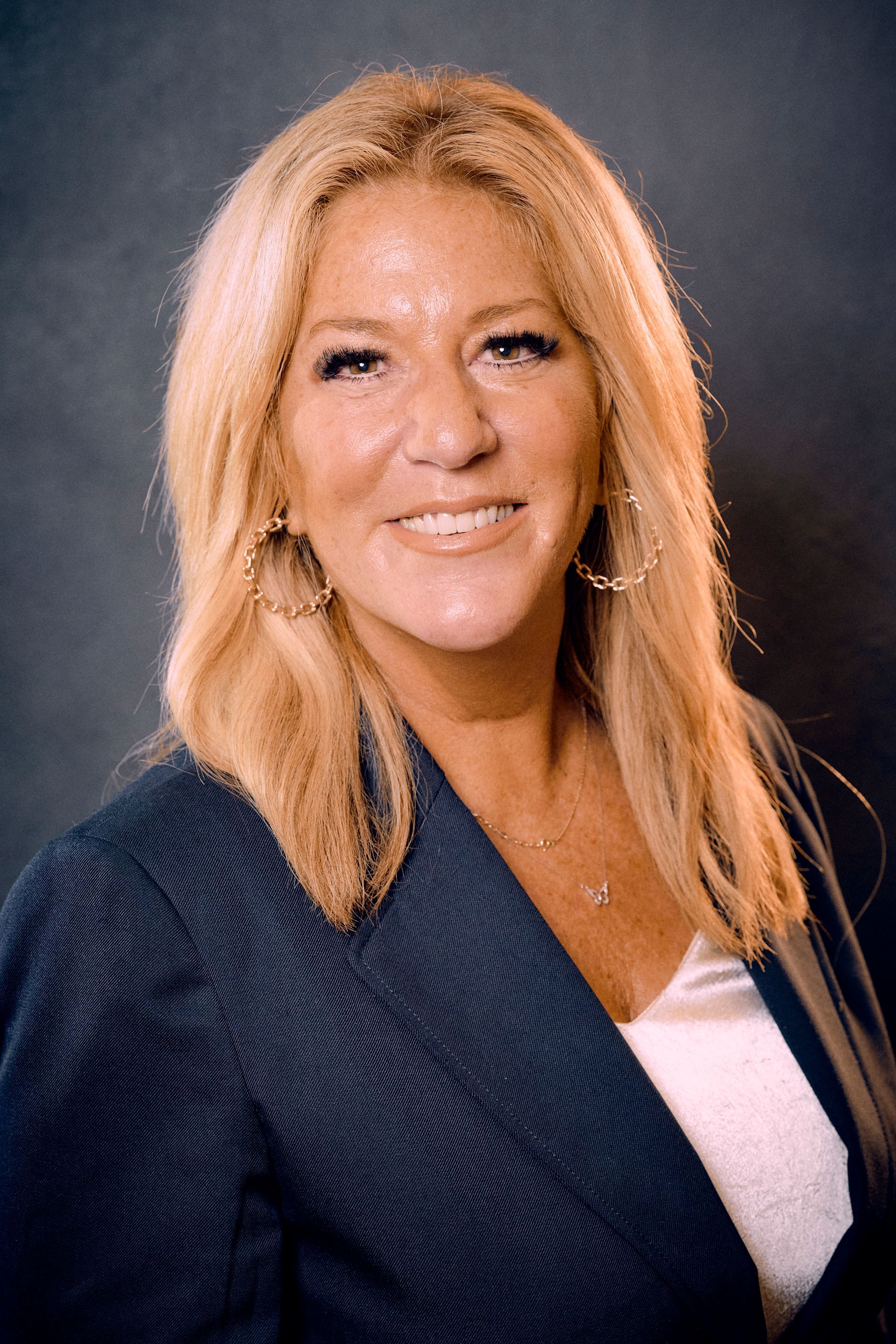 Blonde woman in blue blazer smiling, wearing hoop earrings.