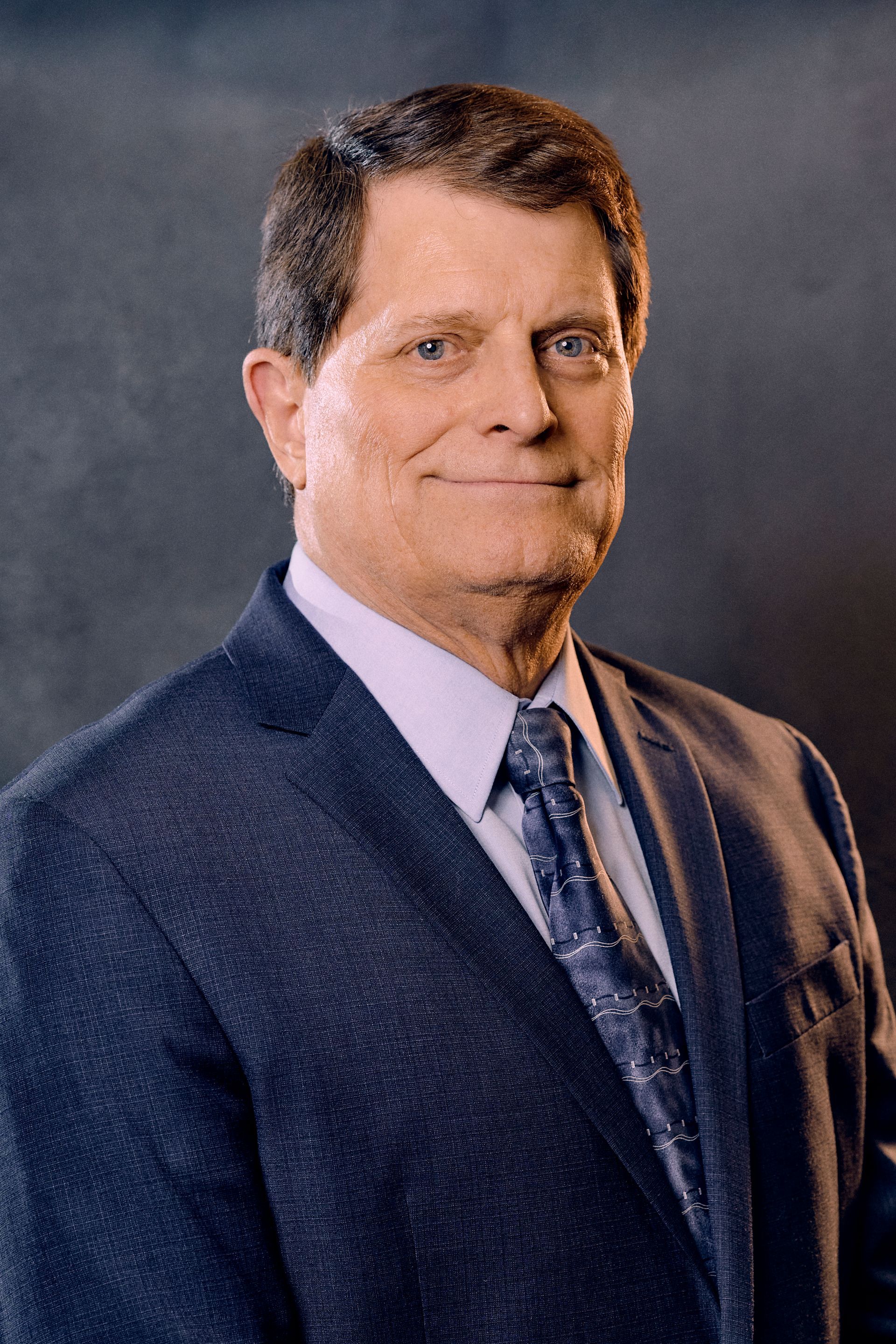 Man in a dark suit smiling; studio portrait against a dark gray backdrop.