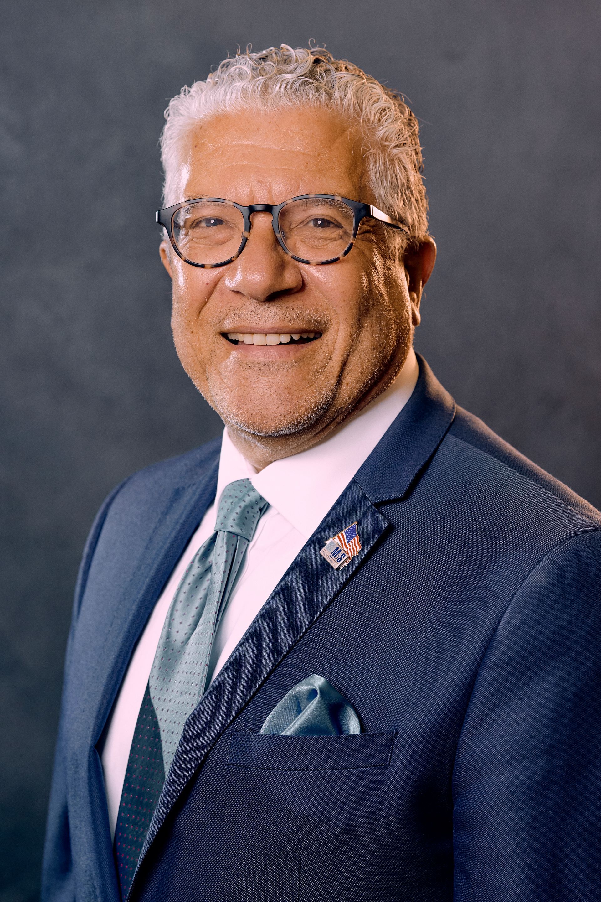A smiling man in a blue suit and glasses. He has gray hair and a teal tie.