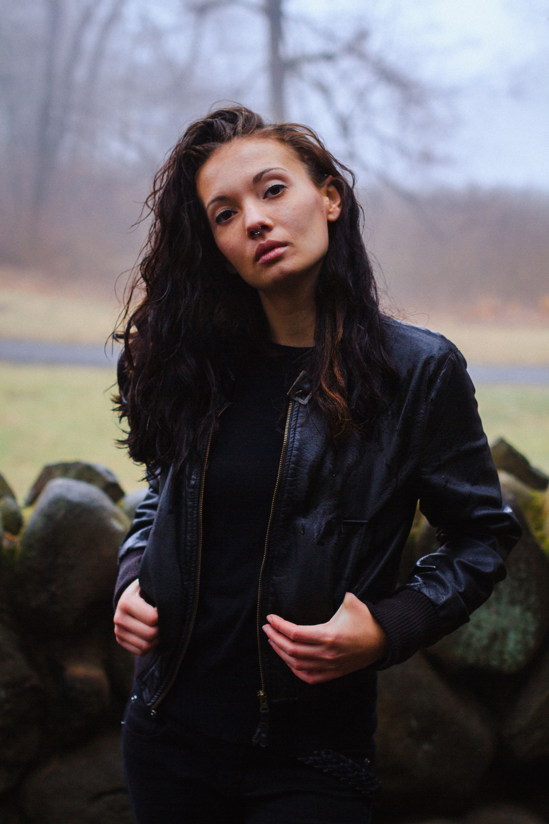 Woman with long dark hair in black jacket stands near a stone wall, looking at the camera.