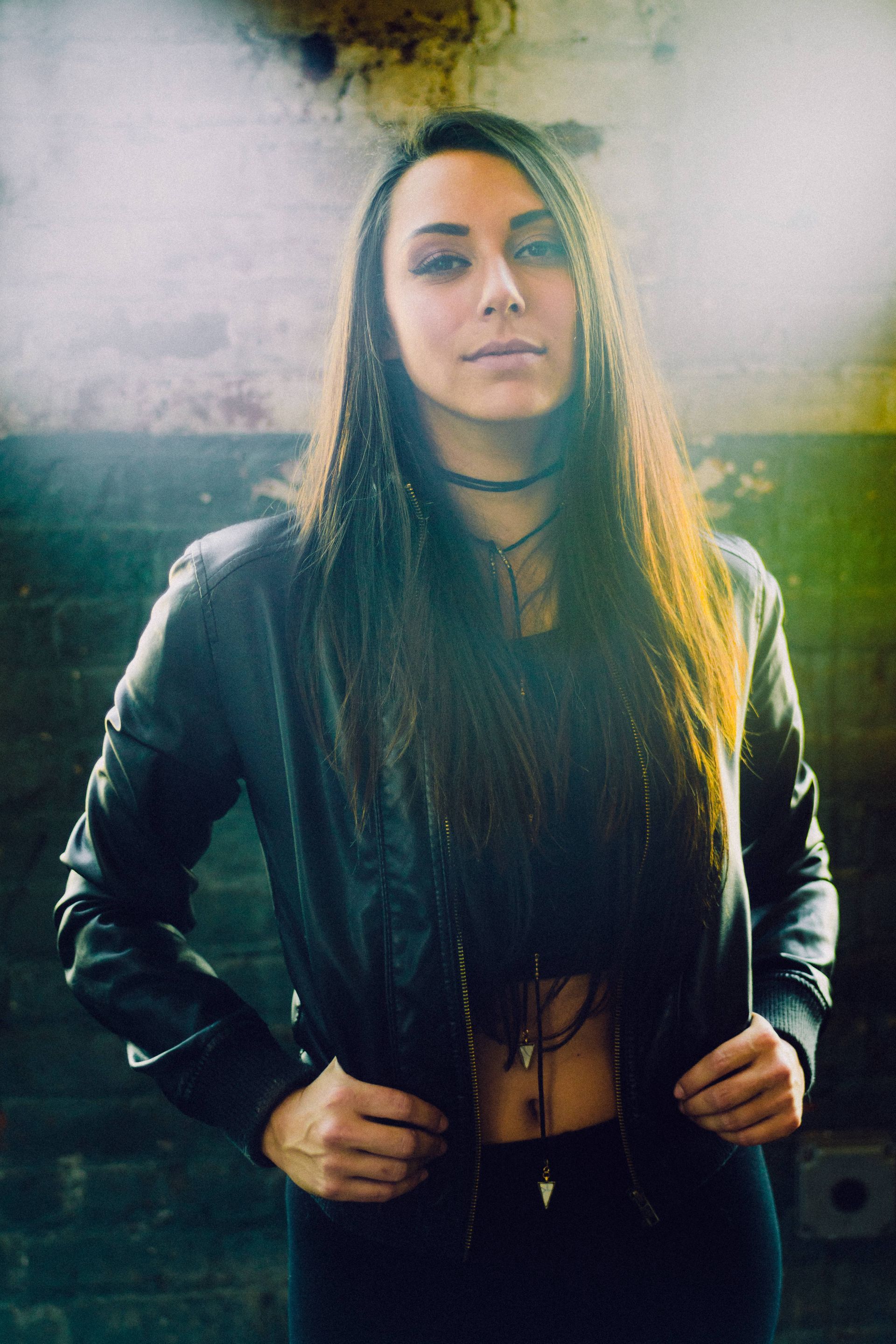 Woman with long brown hair, leather jacket, hands on jacket, standing in front of a brick wall.