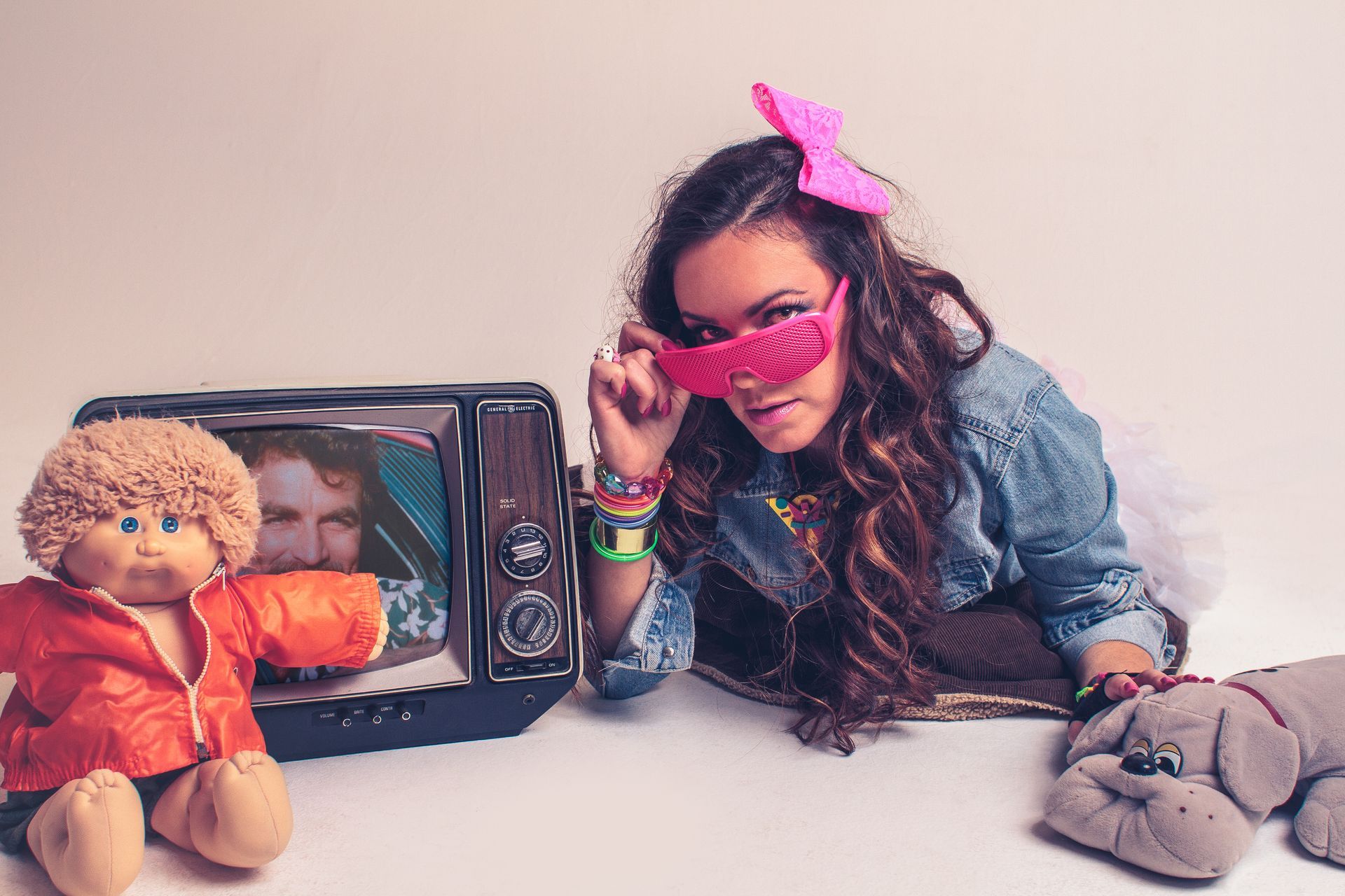 Woman in 80s attire adjusts sunglasses, surrounded by toys and a vintage TV.