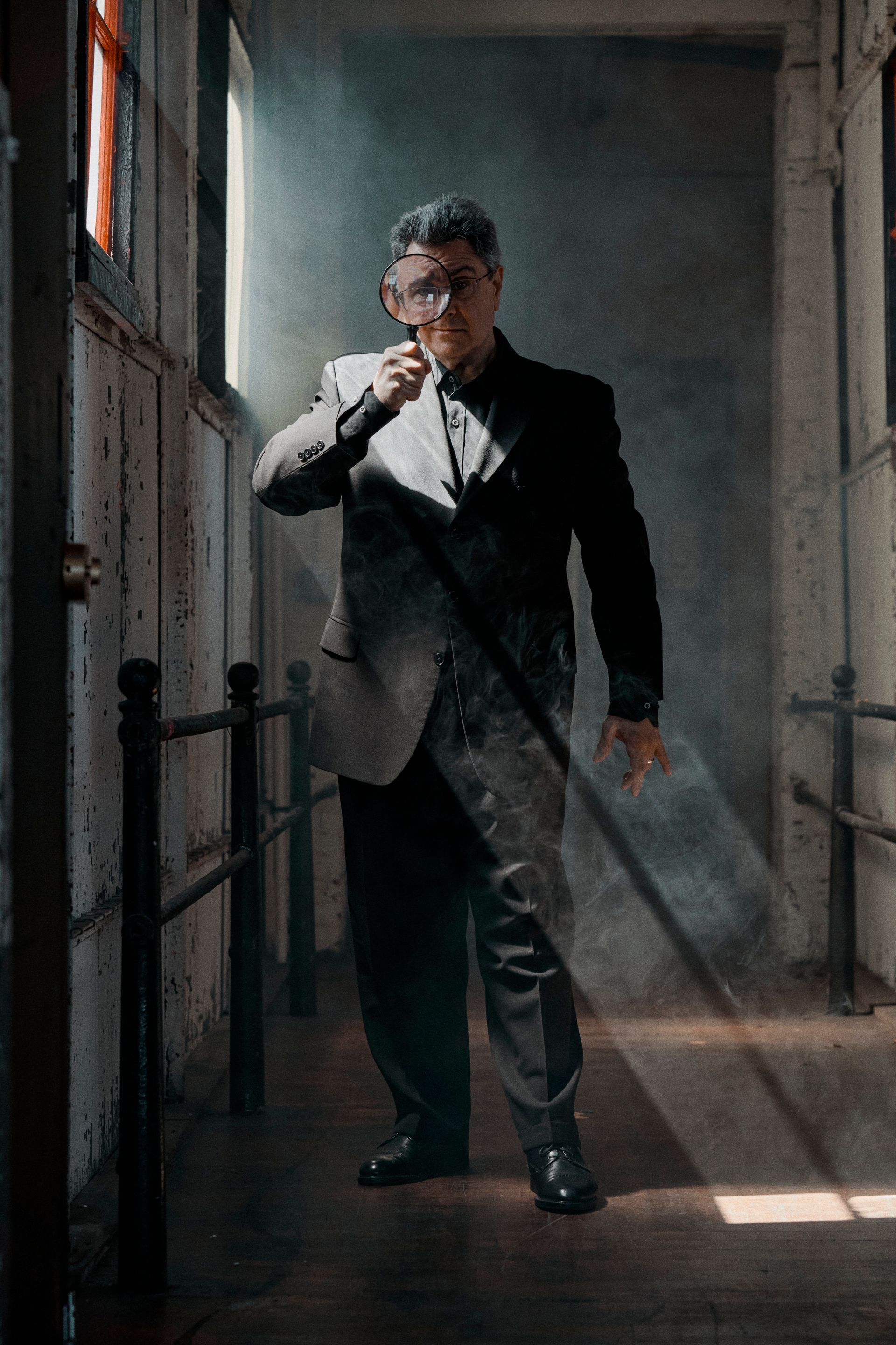 Man in a suit, smoking a cigarette, standing in a dimly lit hallway with sunlight streaming through windows.