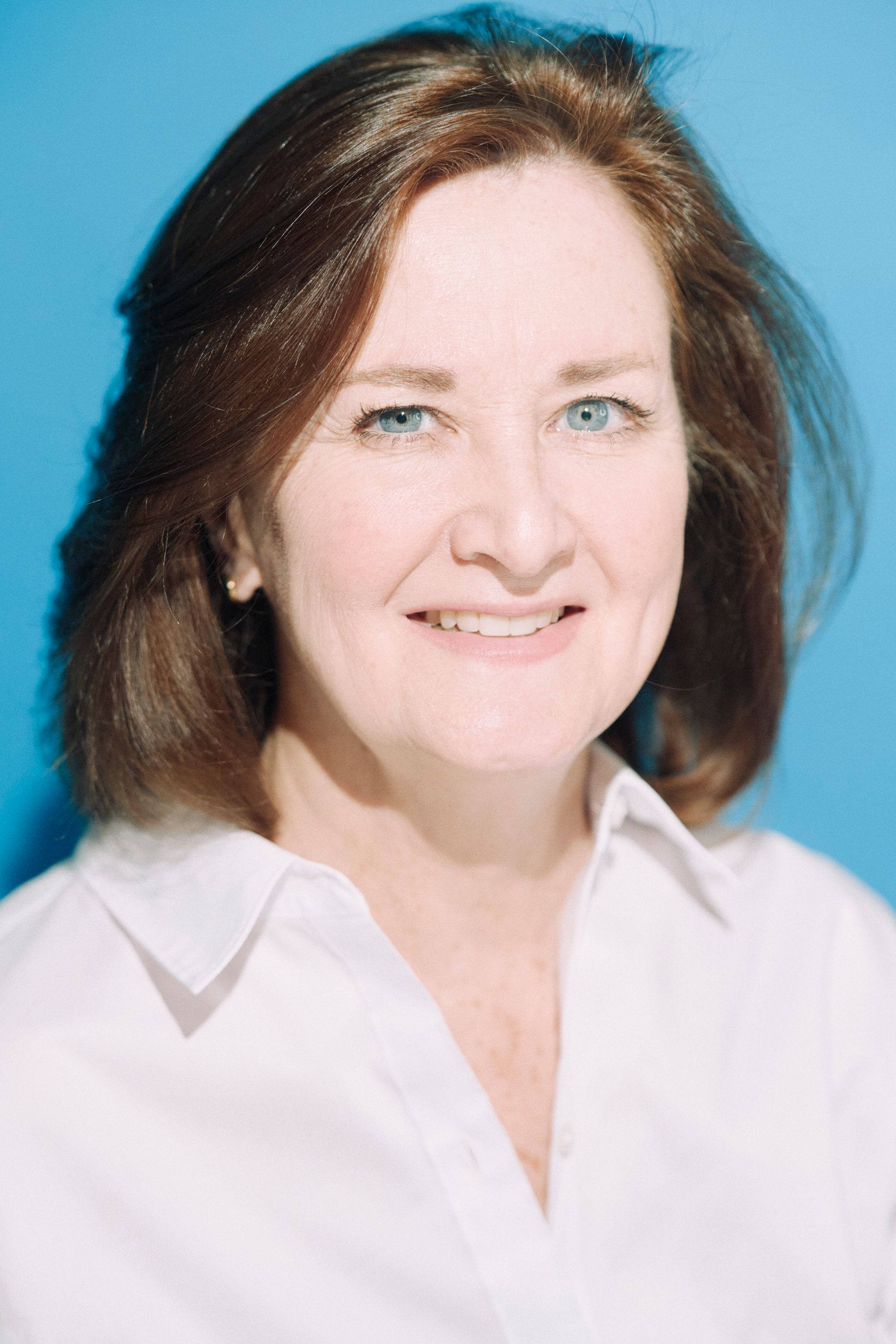 Woman with auburn hair and blue eyes smiles, wearing a white shirt, against a blue backdrop.