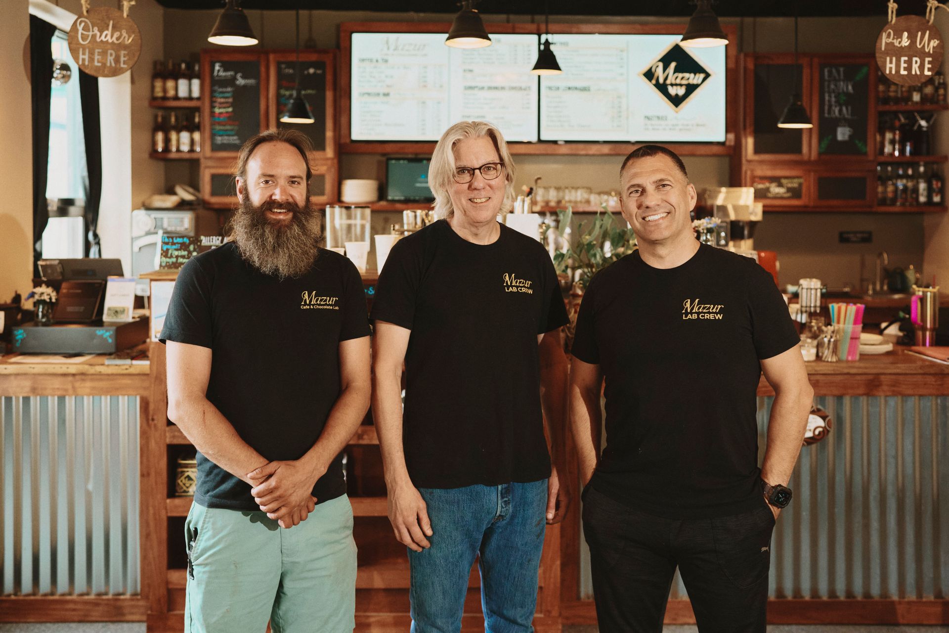Three men in black shirts stand inside a cafe. One has a beard, all smiling. Behind them is a counter.