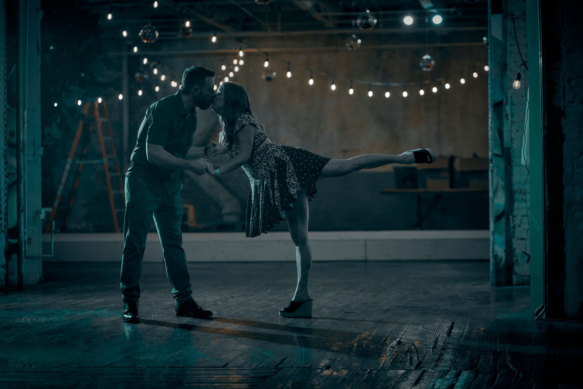 Couple dancing in a dimly lit room. Man kissing woman on the forehead as she extends one leg.