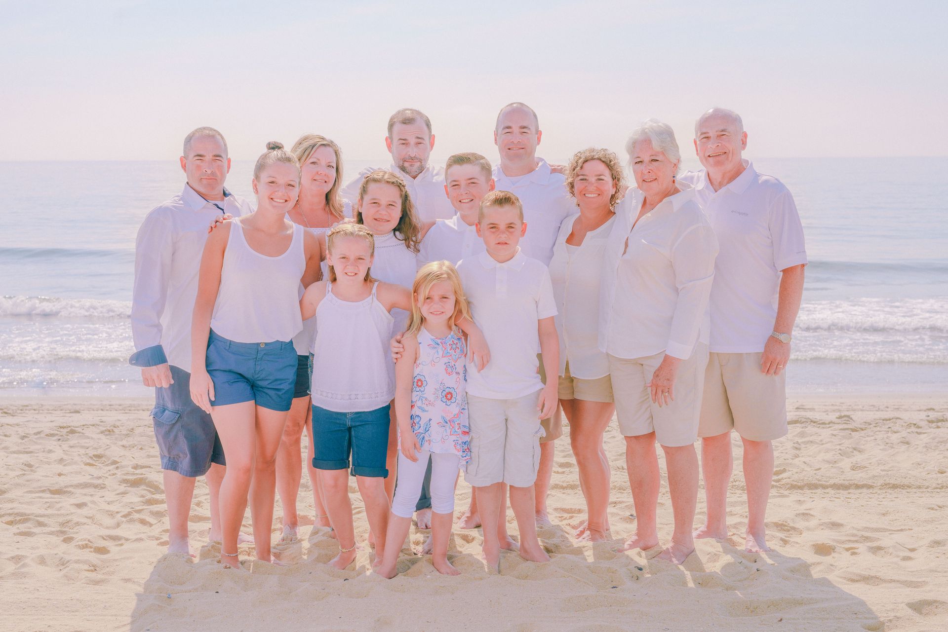 Large family smiling on sandy beach, wearing white and light-colored clothing.