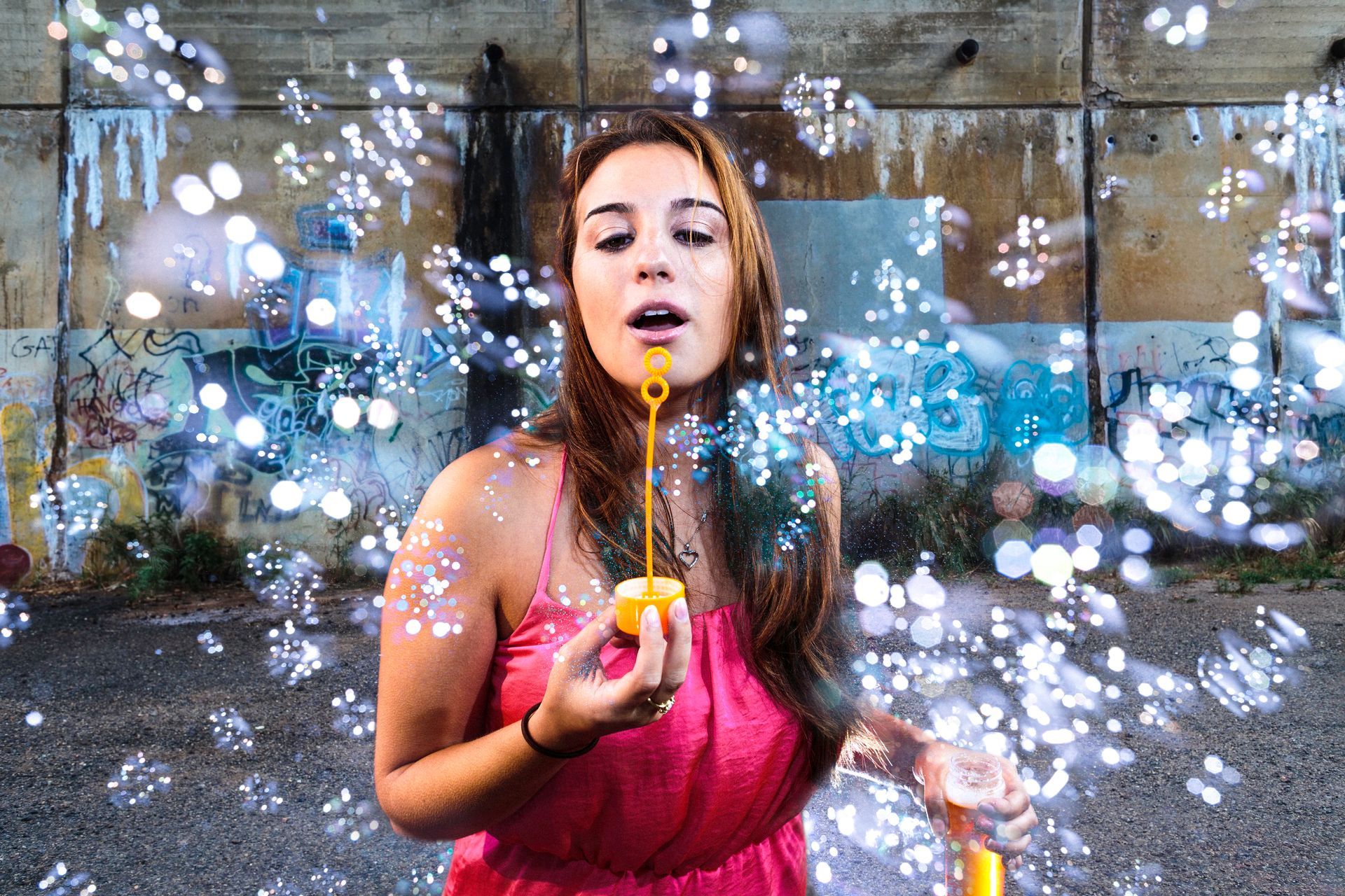 Woman in pink dress blowing bubbles in front of a graffiti-covered wall.
