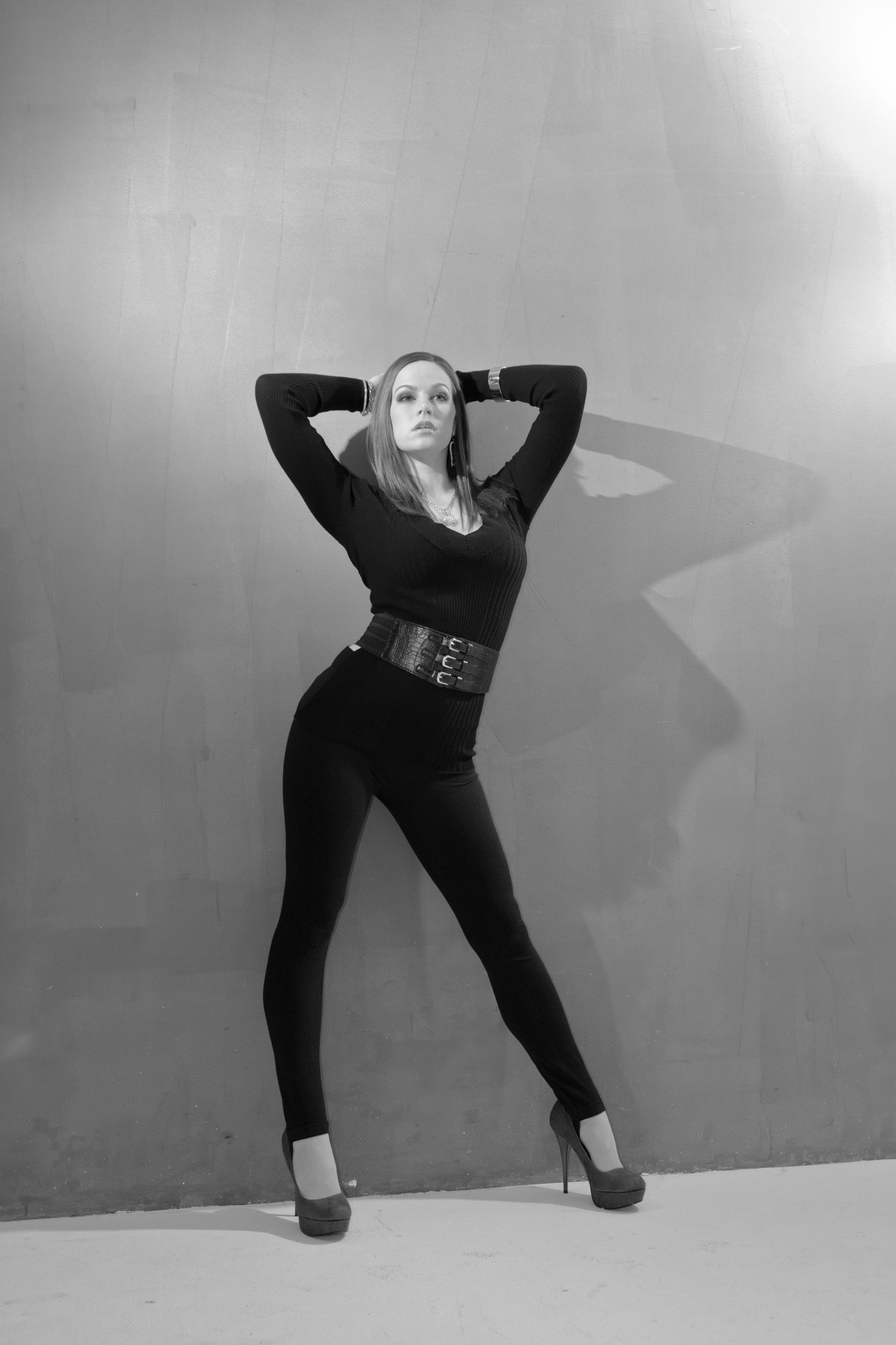 Woman in black outfit, arms over head, leaning against a wall, wearing high heels, black and white photo.