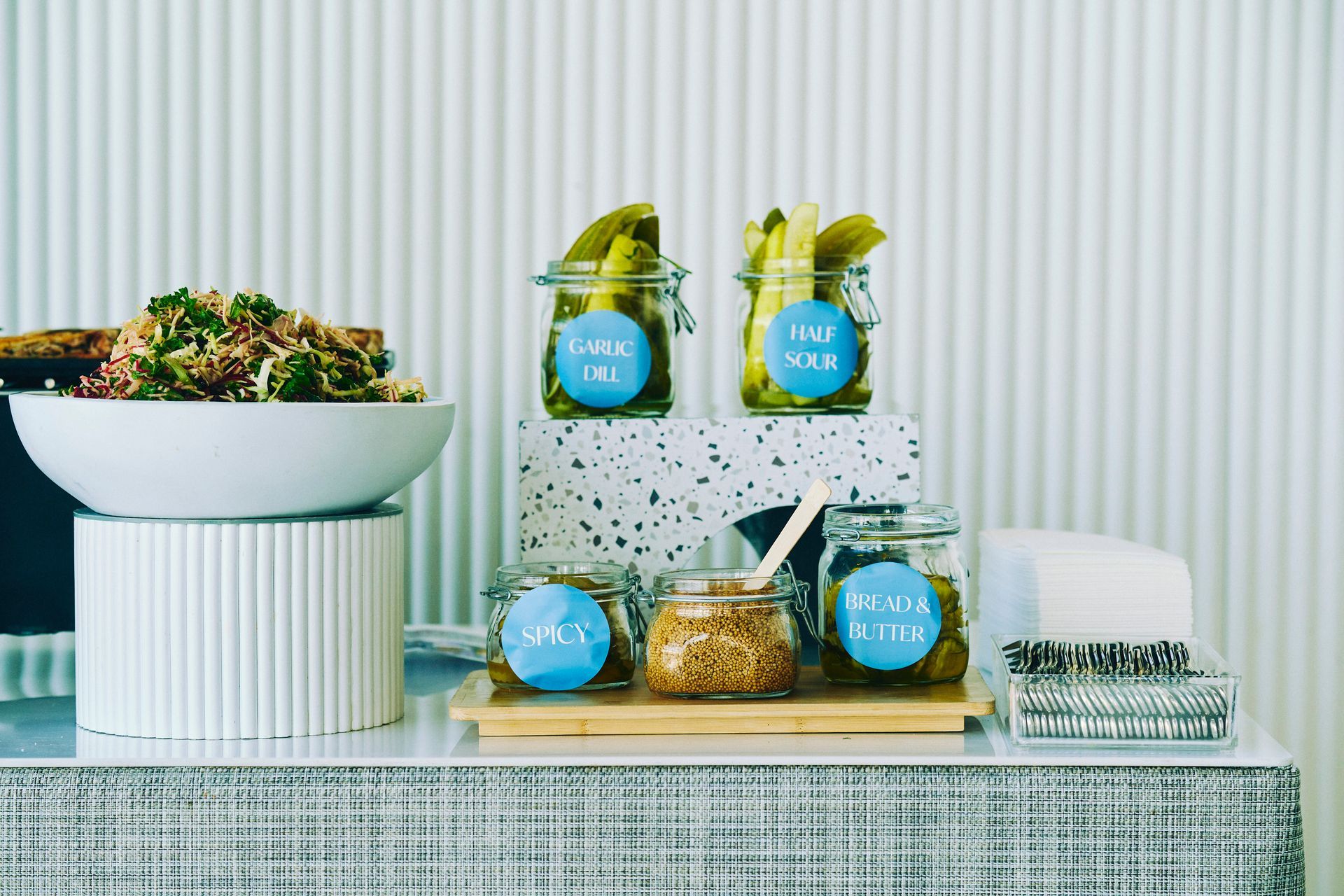 Food display: quinoa salad, pickles, mustard, and napkins on a counter.