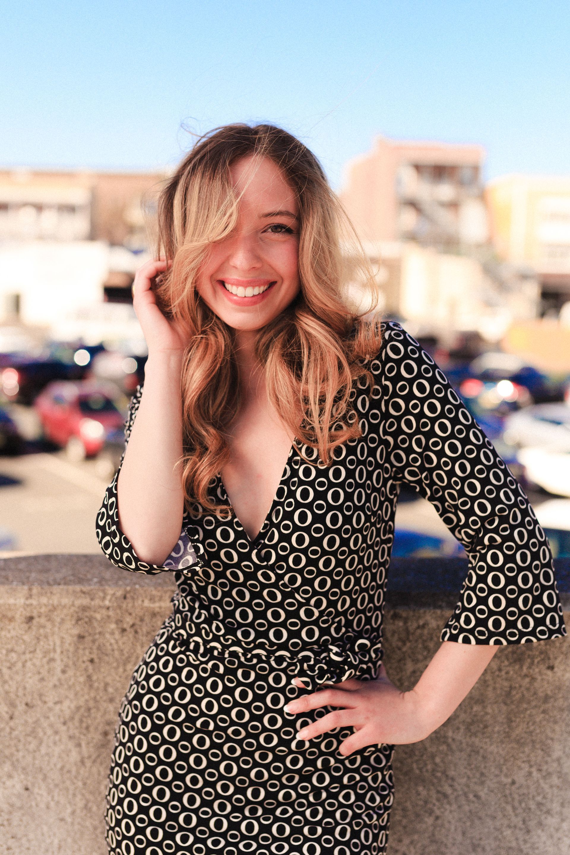 Woman with long wavy hair smiles broadly in a black and white patterned dress outdoors.