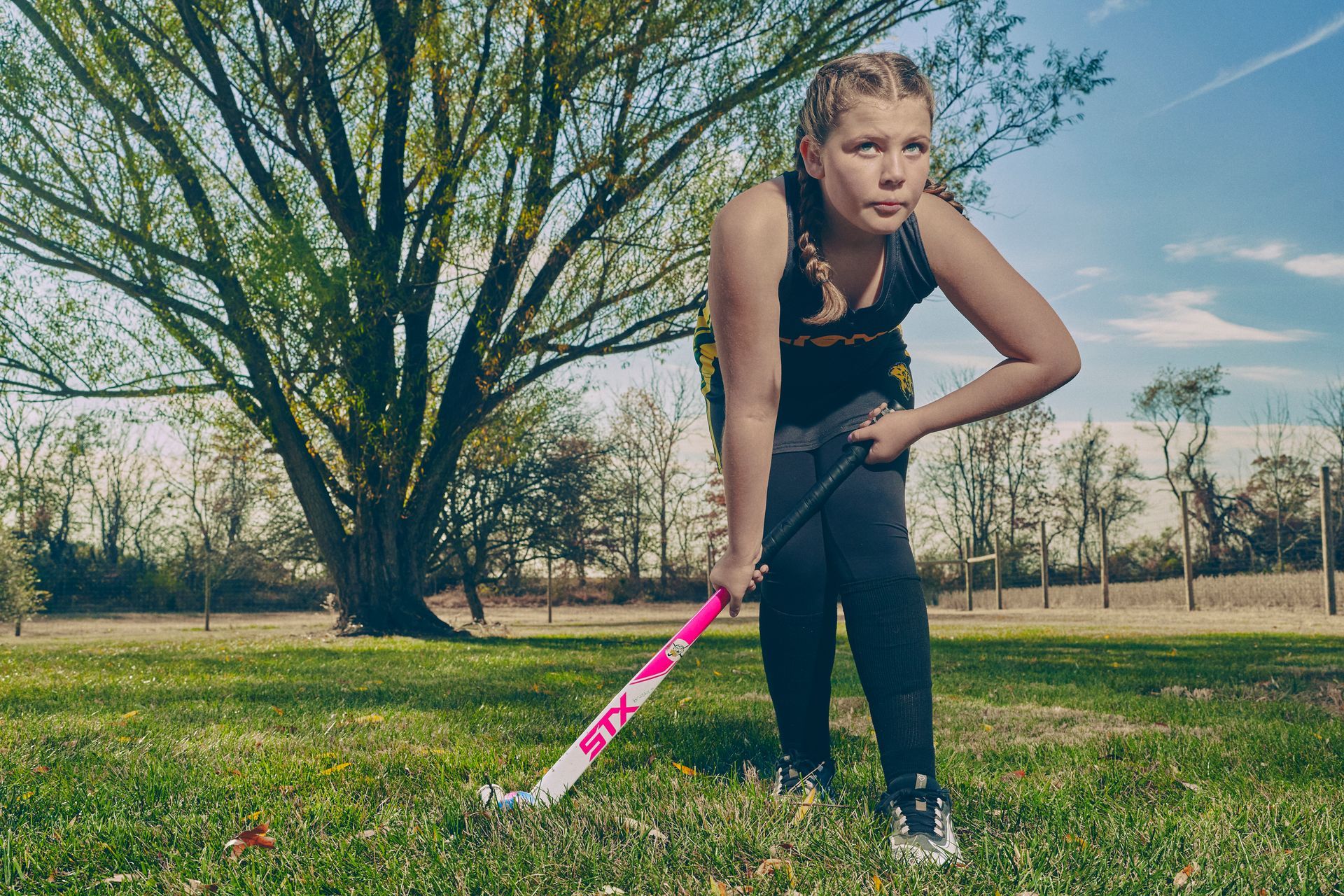 Girl in black outfit on grass, holding a pink field hockey stick, tree in background.