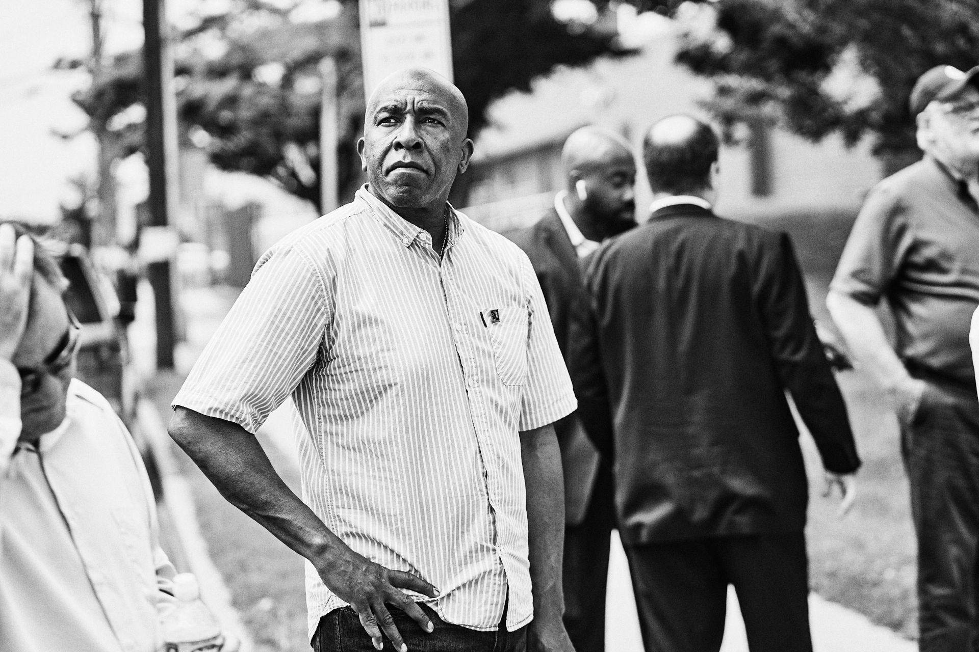 Black and white photo of a Black man in a striped shirt looking off to the side; other men stand nearby.