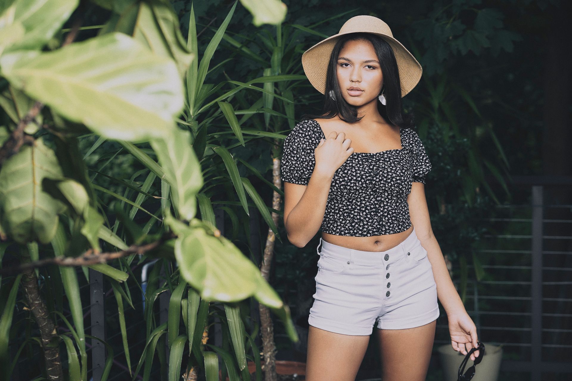 Woman in straw hat, floral top, and shorts posing outdoors by greenery.