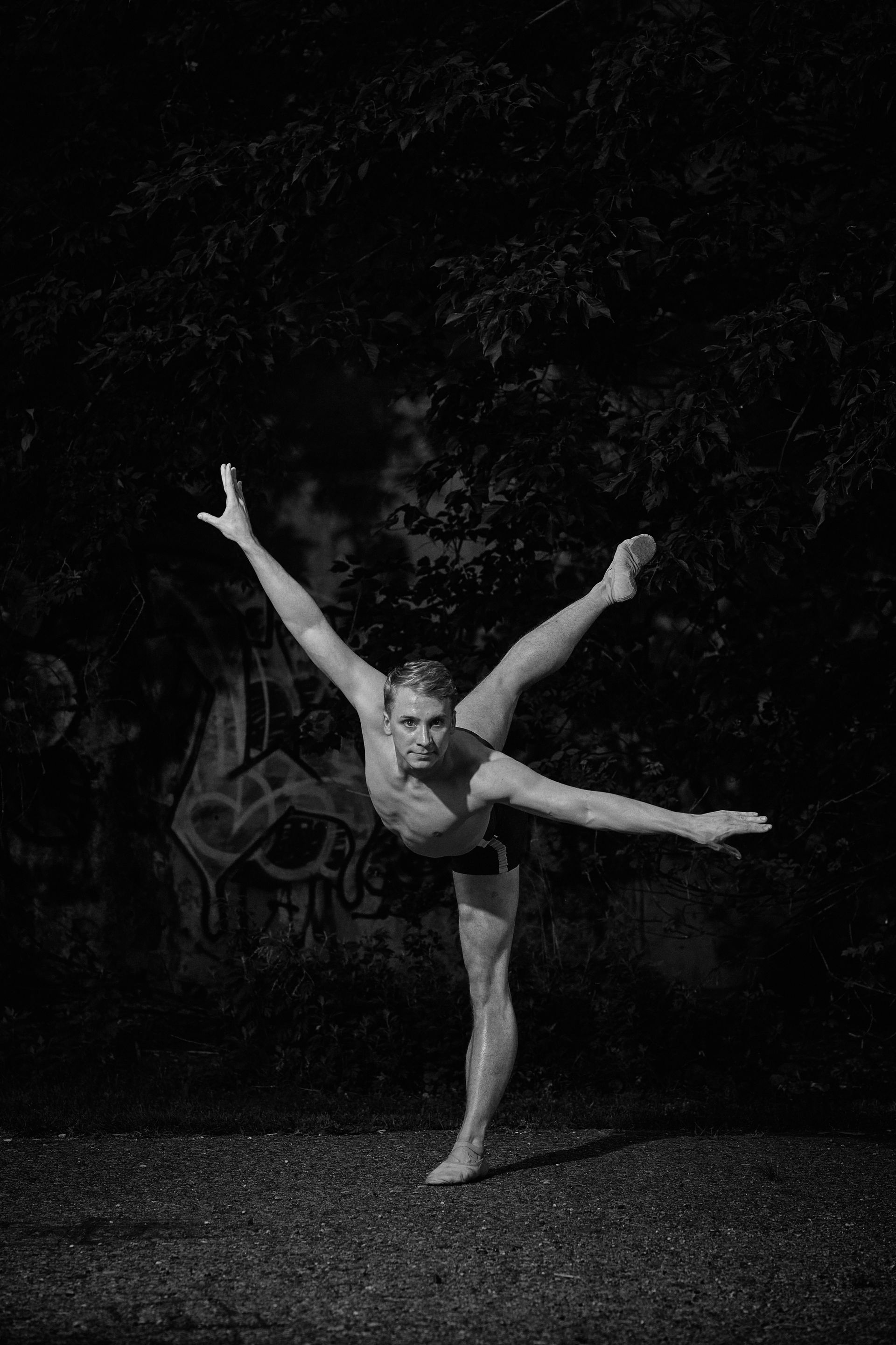 Ballet dancer leaping mid-air, arms outstretched, under a bridge near green trees.
