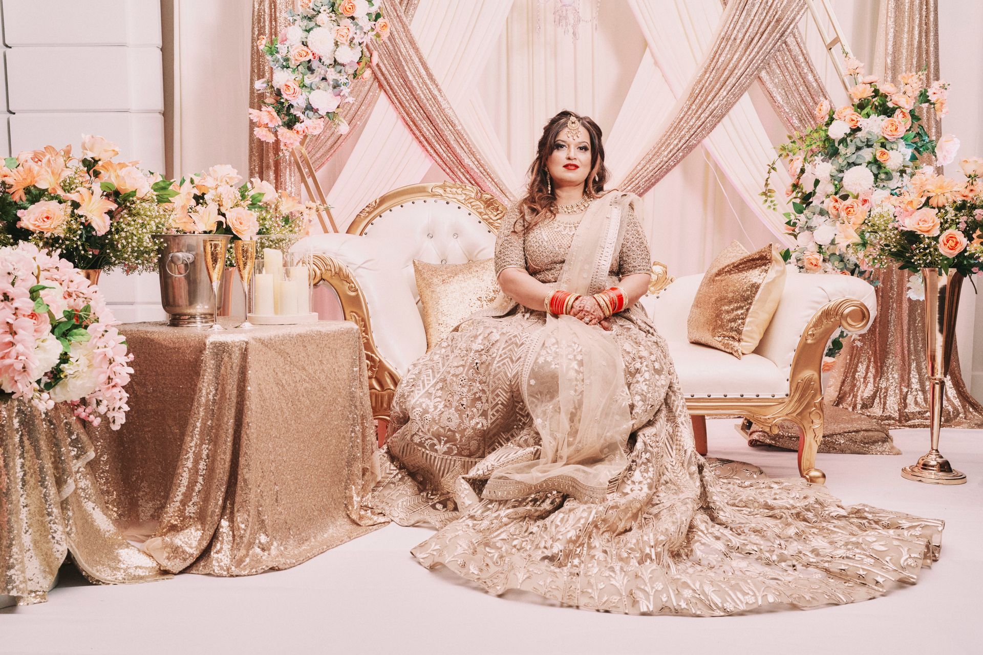 Woman in gold sequin dress sits on ornate sofa, posing for a photo at a decorated event.