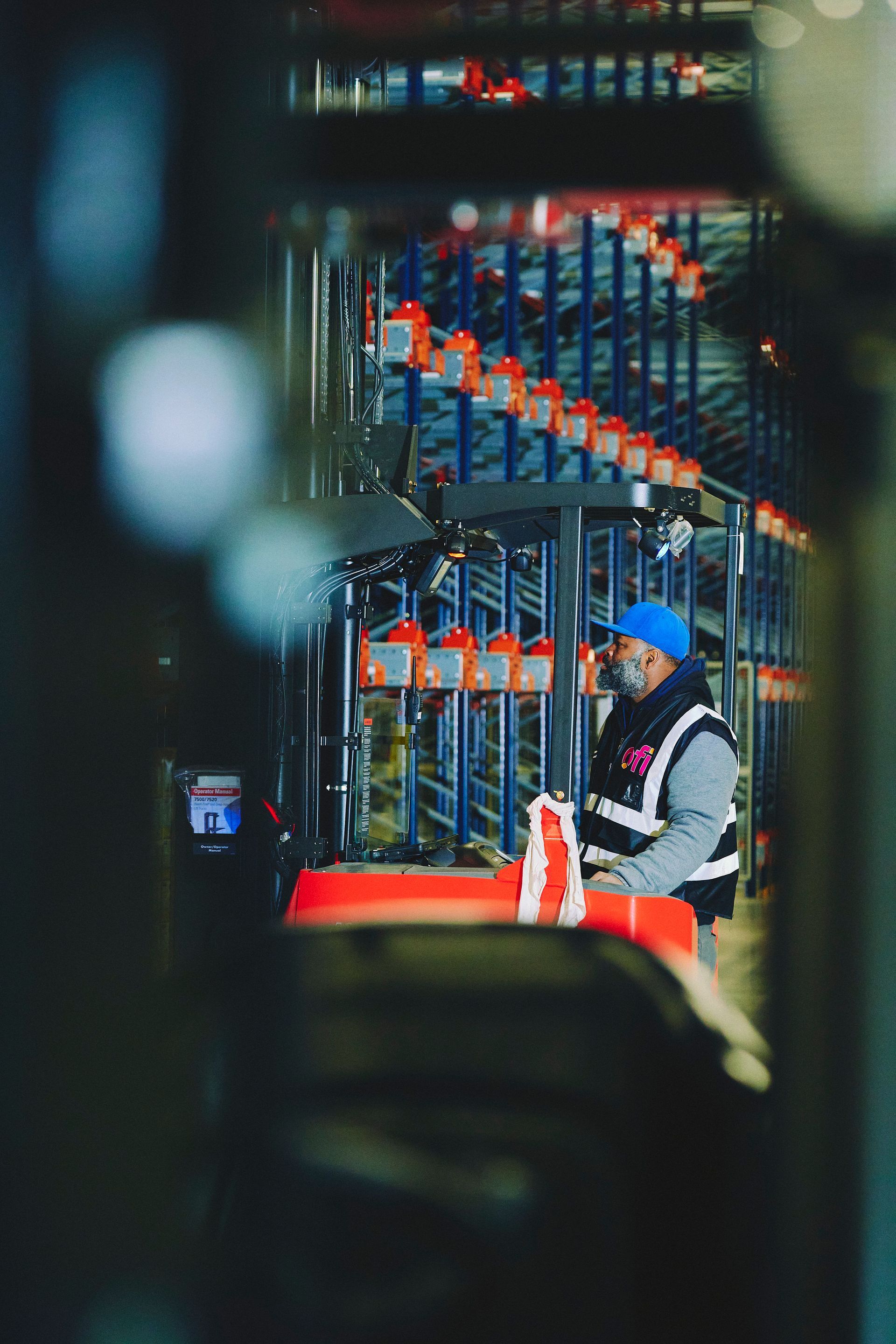 Warehouse worker operating machinery in front of a tall shelving system. The worker is wearing a blue hat and safety vest.
