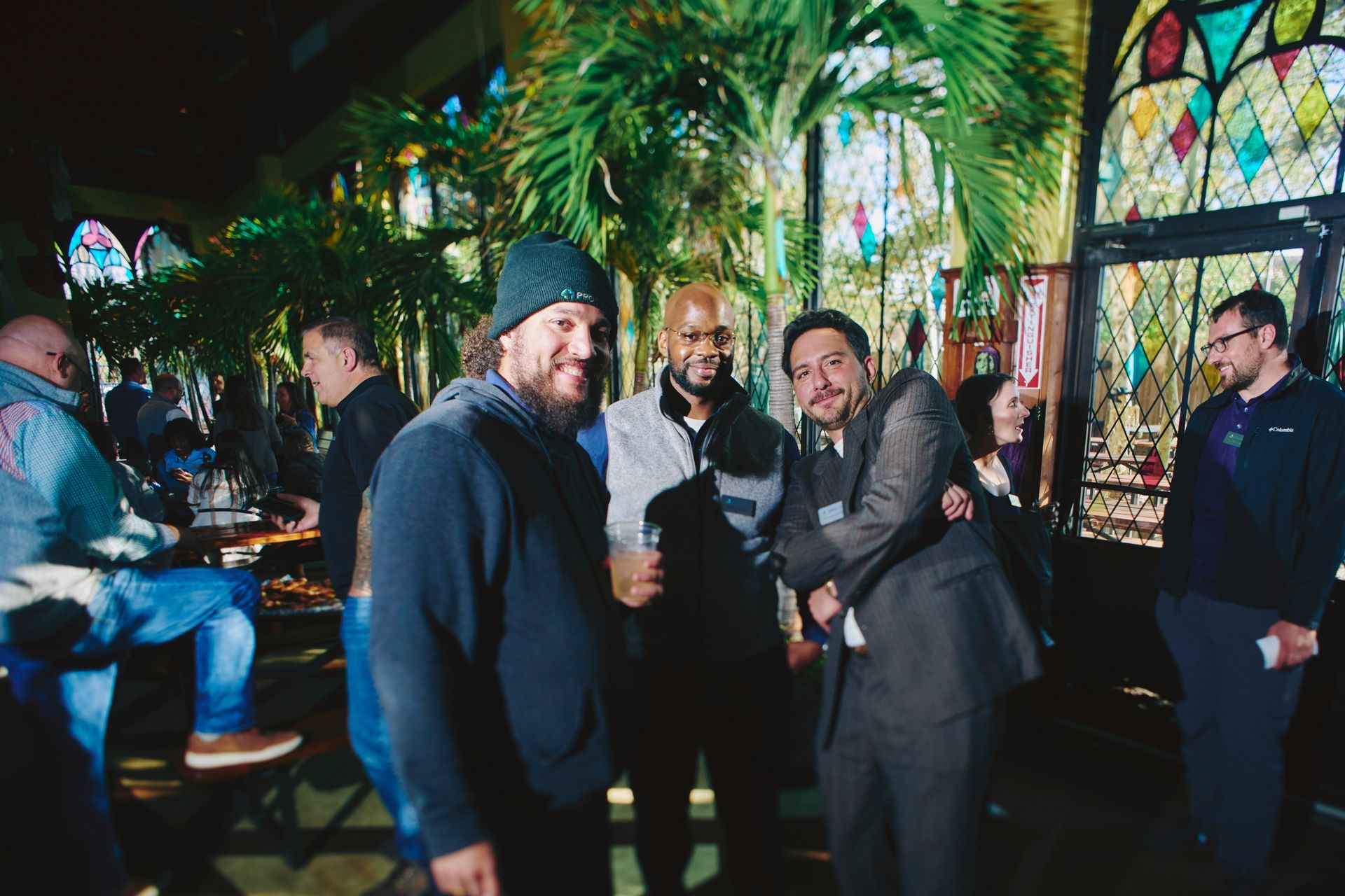 Three men smiling, posing inside a building with stained glass windows and palm trees. People in the background.