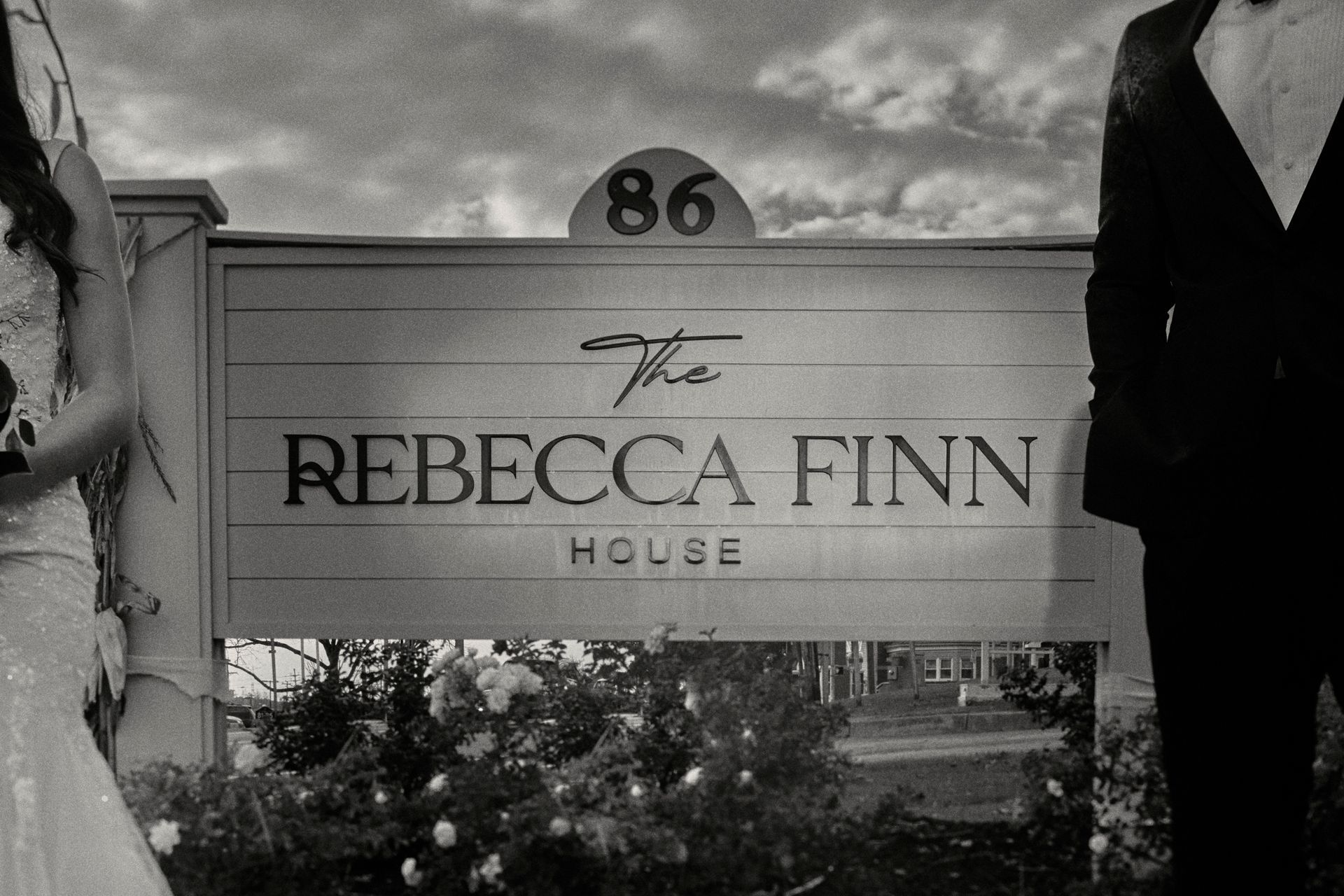 Sign for The Rebecca Finn House, 86. A couple in formal wear stands nearby; black and white.