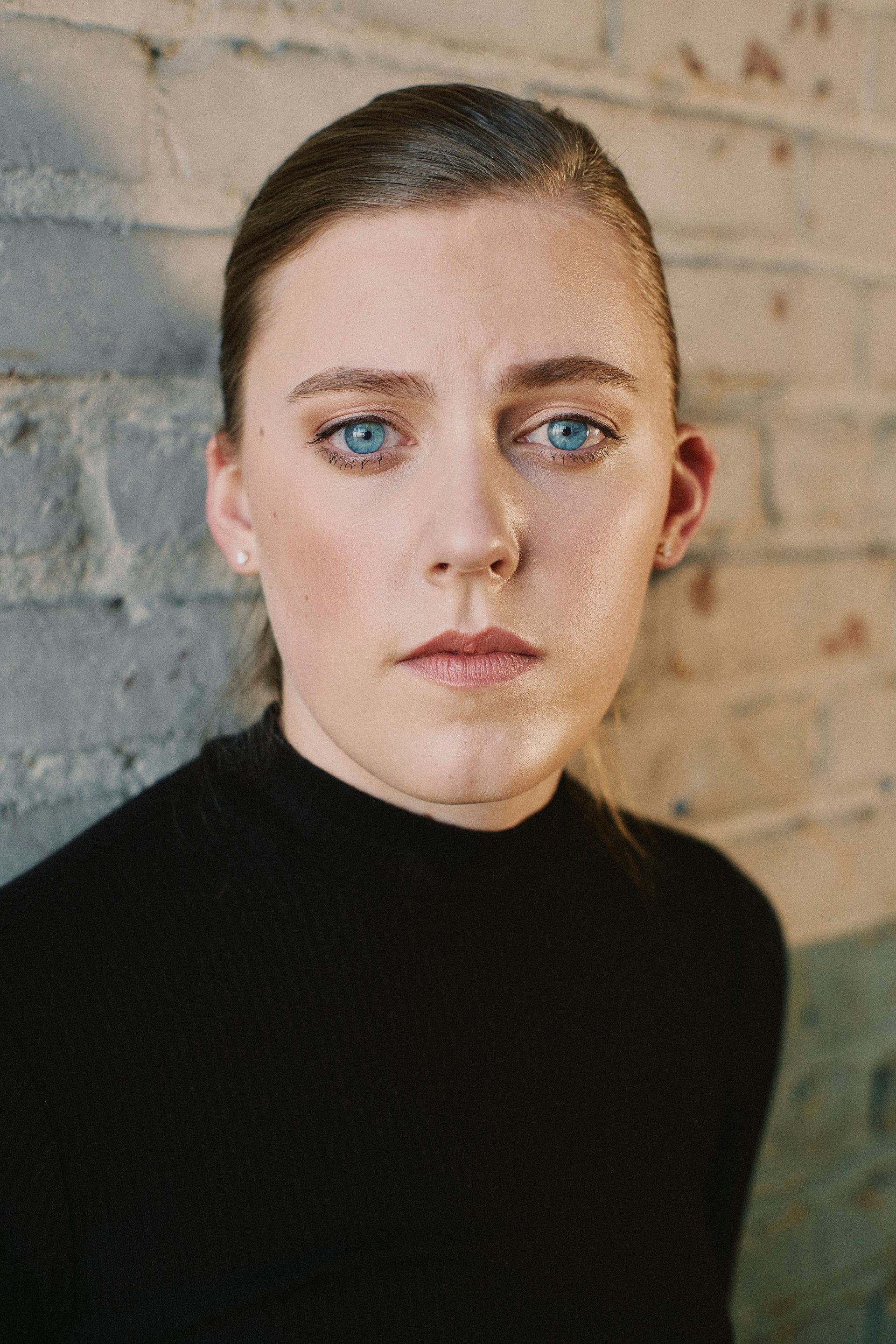 Woman with blue eyes, dark hair, black top, leaning against a brick wall, serious expression.