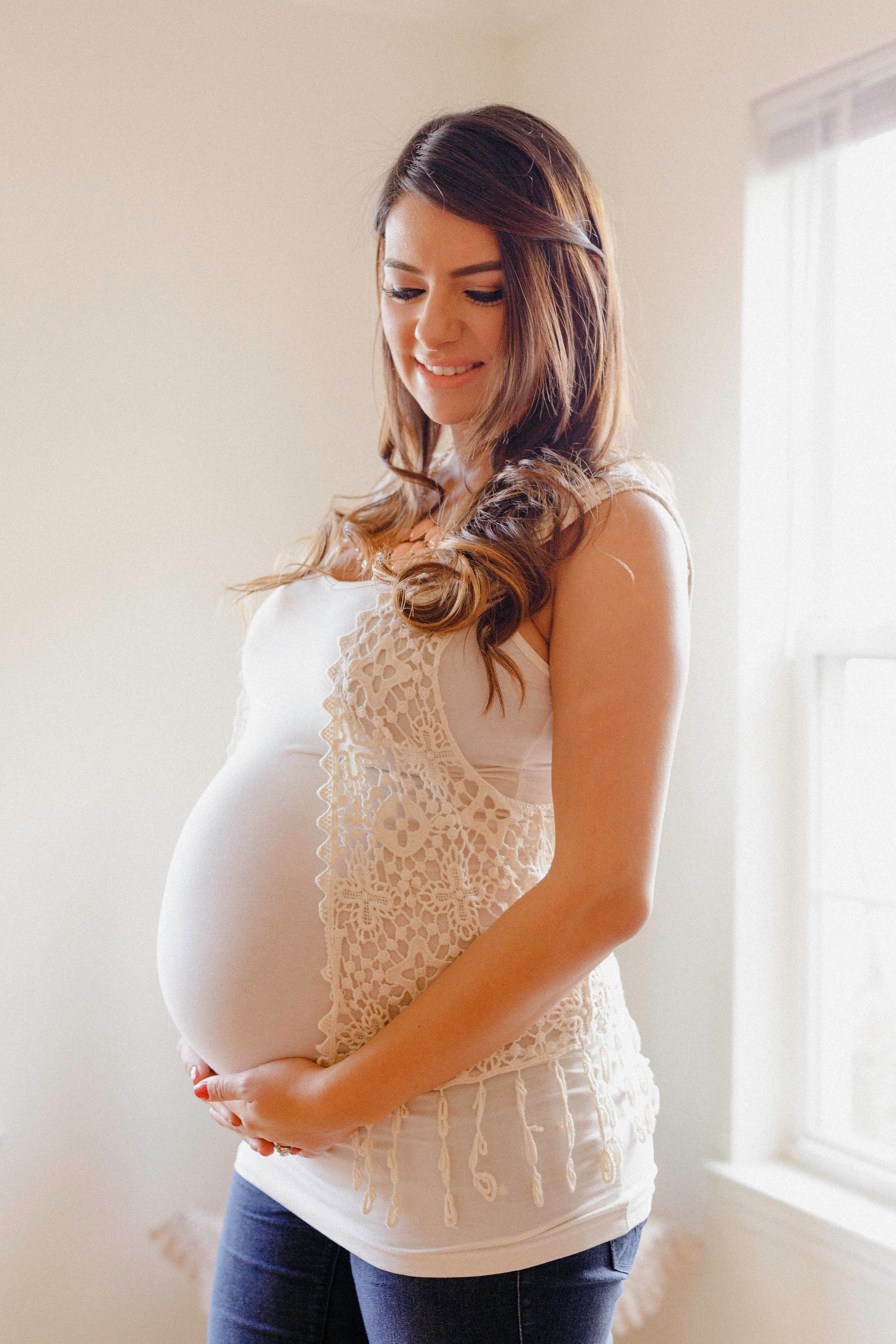 Pregnant woman in a white top and jeans, hands on her belly, smiling near a window.