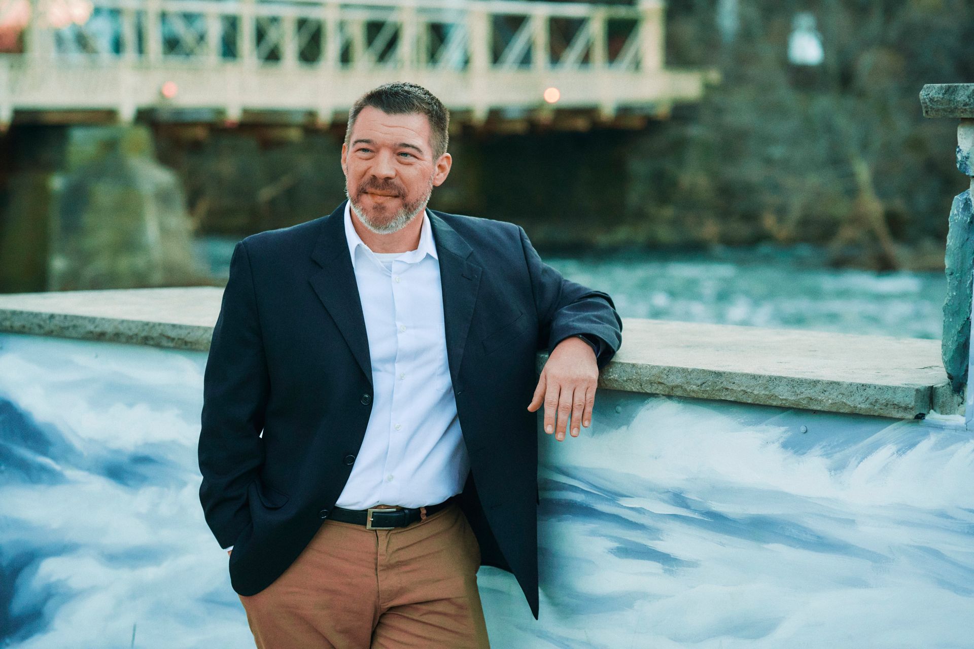 A man in a suit is leaning against a wall next to a river.