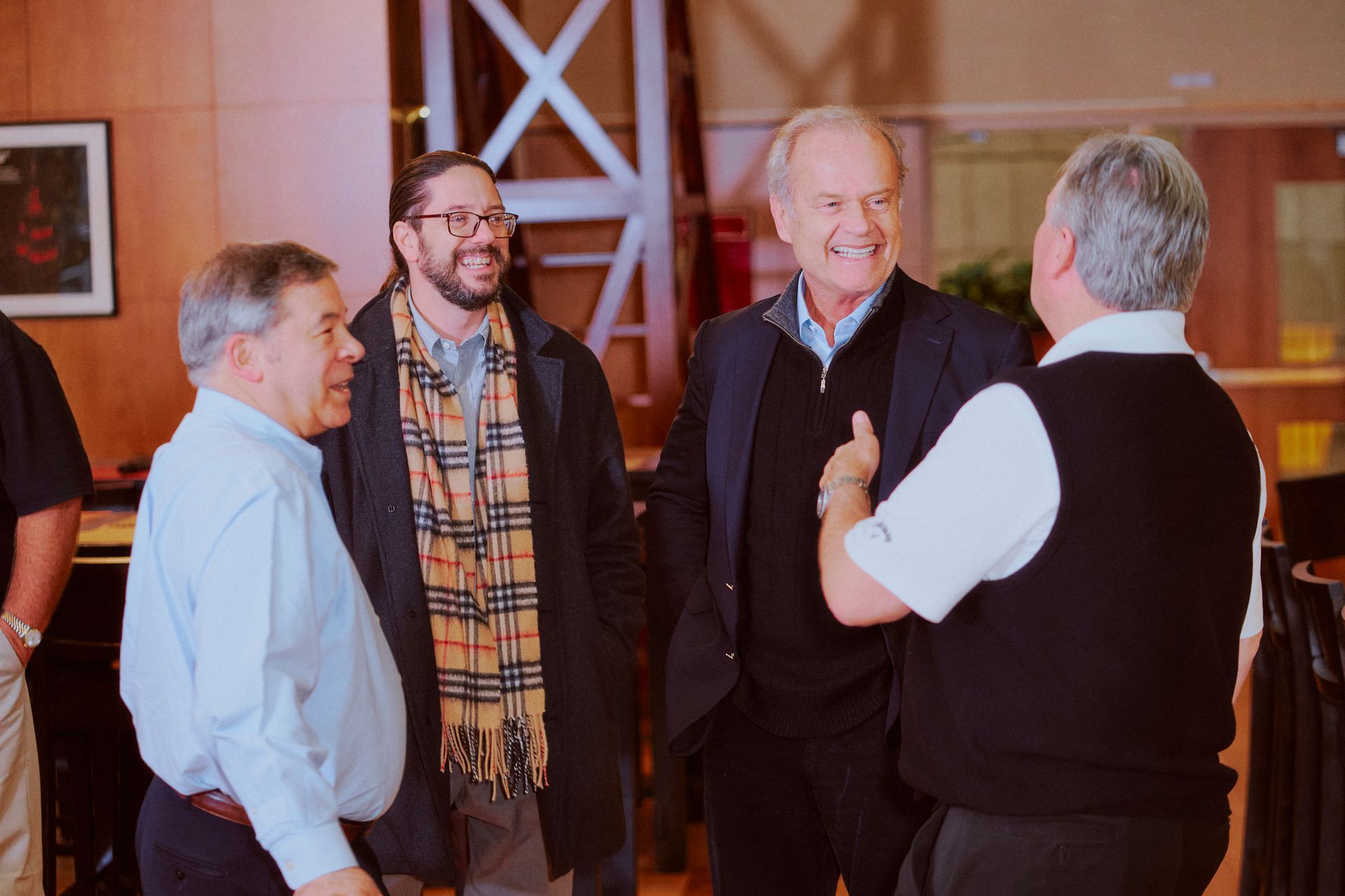 Four men conversing in a well-lit room. Two smile, one in a jacket. One in a scarf.