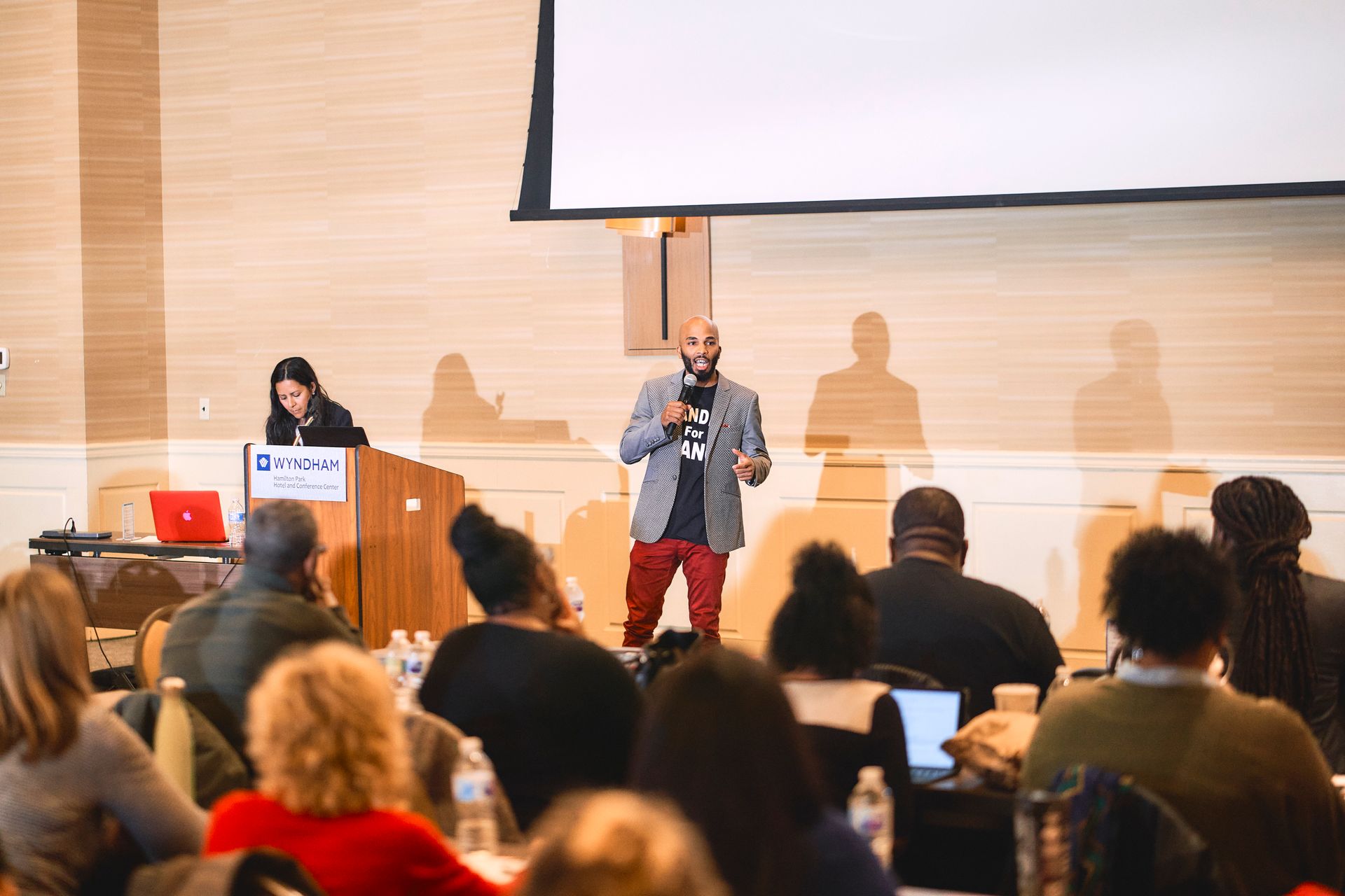 Man speaking at conference, red pants, holding a mic, in front of seated audience and a woman at a desk.
