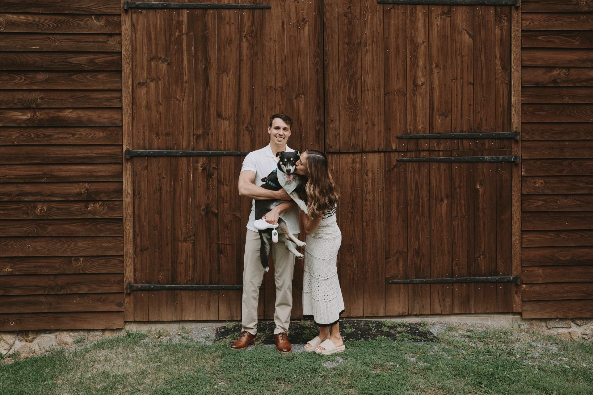 A man and woman are holding a dog in front of a wooden building.