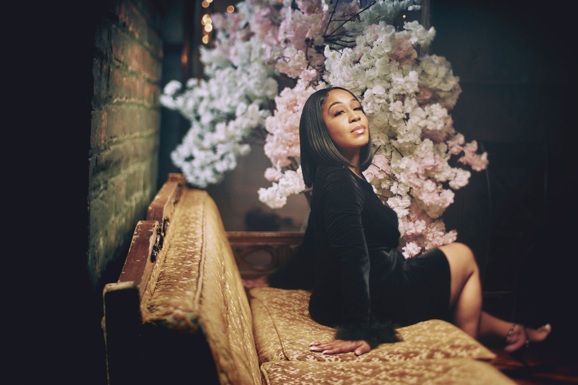 Woman in black dress on gold couch, smiles, under pastel flowers in dark room.