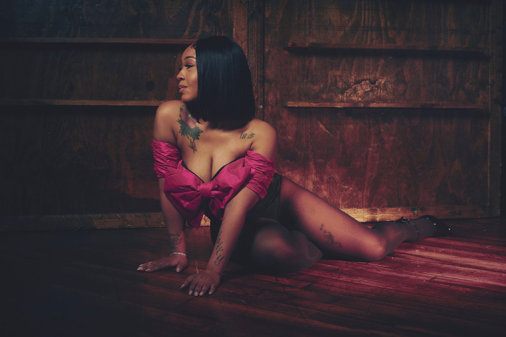 Woman in pink off-the-shoulder top, sitting on floor, dark room, looking to side.