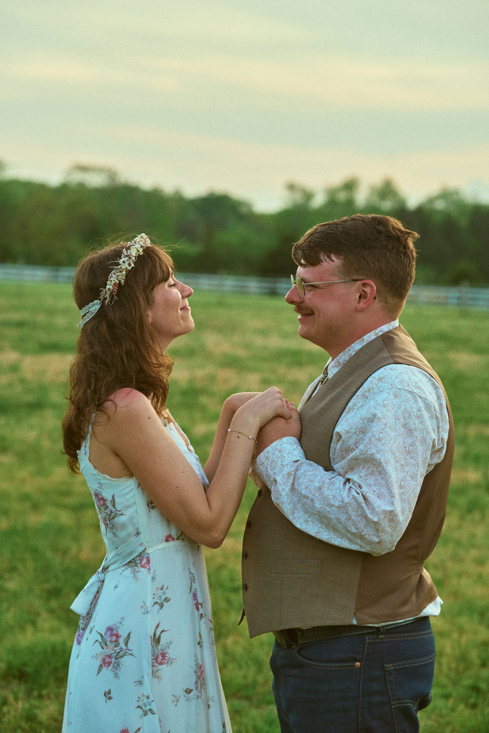 Couple holding hands, smiling at each other in a green field. Woman wearing a floral dress, man with a vest.
