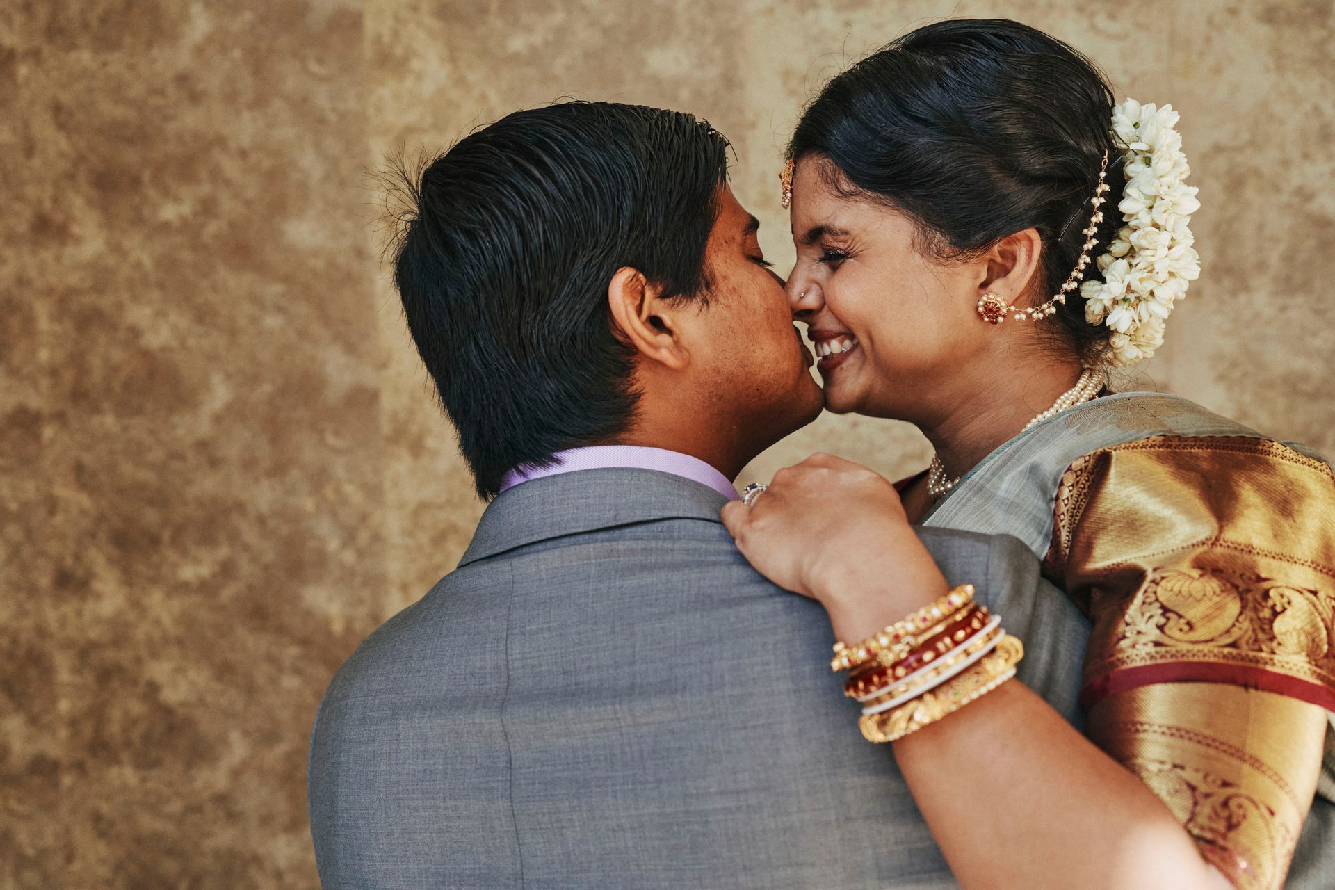 Couple kissing, woman in saree, man in suit, smiling, against a textured wall.