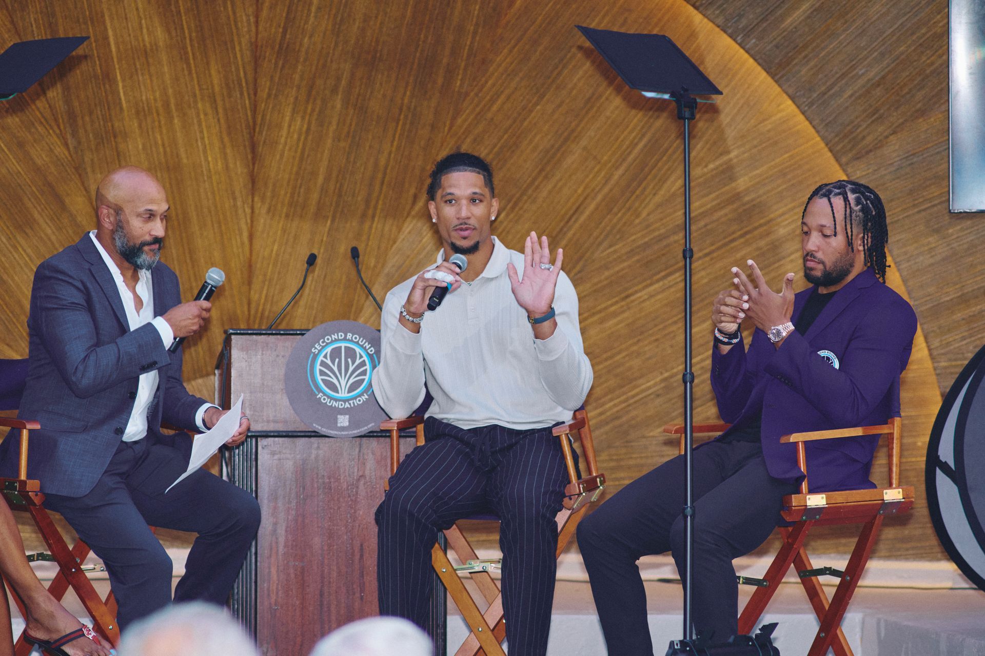 Three men on stage at a panel. One speaks into a microphone, gesturing with his hand. Another listens and the third holds a mic and listens.
