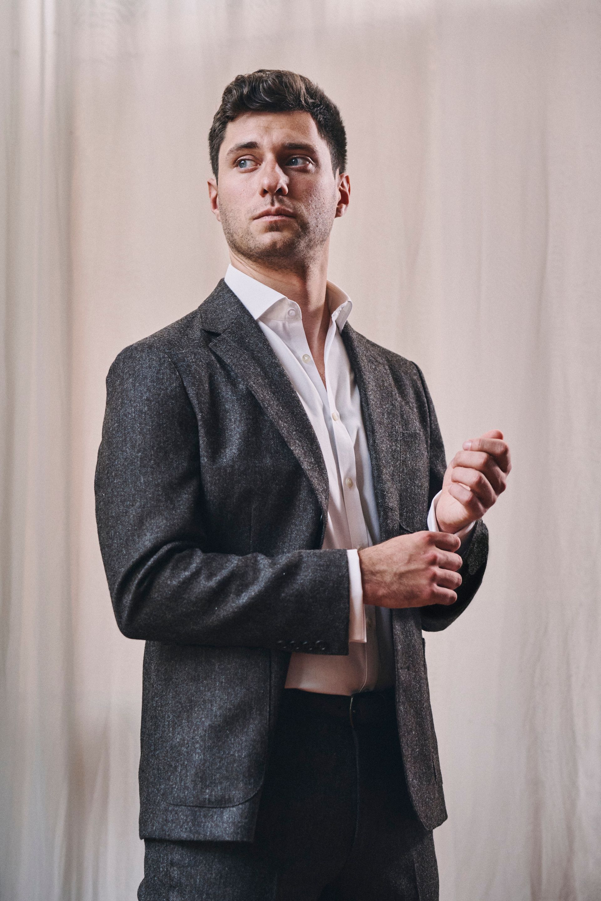 Man in a sparkly gray suit and white shirt, adjusting his cuff. Soft, neutral background.