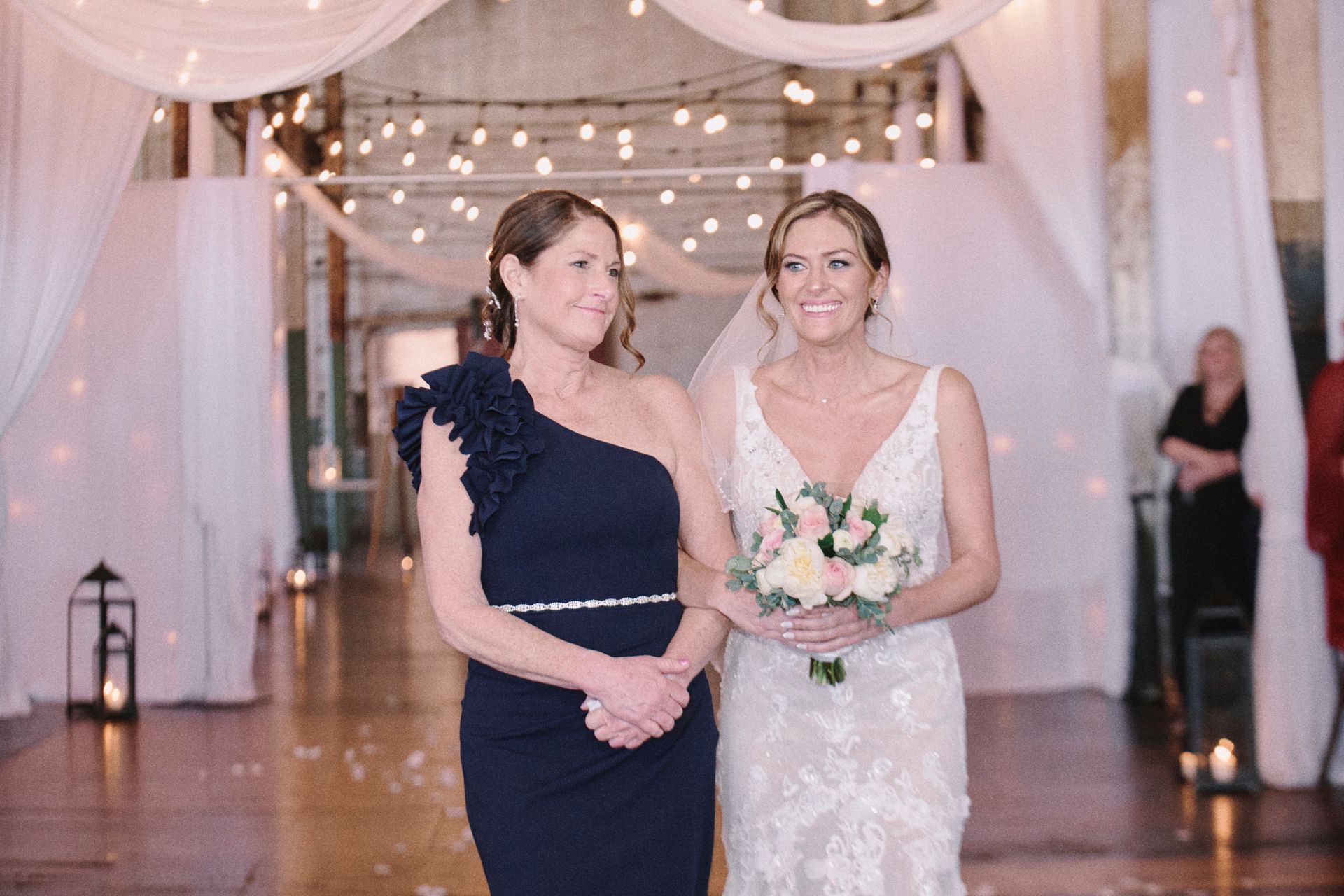 Bride and woman in navy dress walk down an aisle, decorated with draping white fabric and lights.